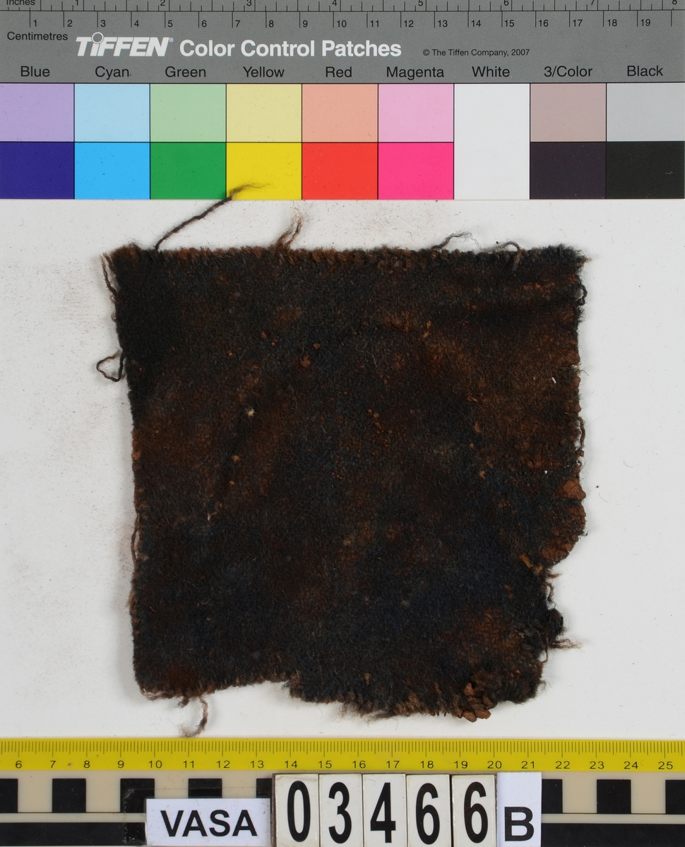 Textil. 13 tygfragment uppdelade på fyndnummer 03466a-d.
Fnr 03466a består av 10 fragment av ull vävda i tuskaft varav det största är ihopsytt av flera delar. Har varit ett par byxor med lucka fram. Originalfärgen kan ha varit blå då rester av blå färg syns på insidan av två av bitarna på de delar där sömmen varit och tyget originalyta bevarats bättre. På en av dessa bitar som tolkats som en del av en ficka på byxans vänstra sida finns rester av en tvåtrådig, s-tvinnad, z-spunnen tråd i sömmen. Byxans tyg har varit mer valkad på insidan än på utsidan. På det största fragmentet framsida syns två knapphål i linningen, och rester efter ytterligare två hål. Det kan därför konstateras att det ursprungligen har varit två knapphål på var sida på linningens mittdel där sidodelarna av linningen knäppts eller knutits fast. Vid ena knapphålet syns något som kan vara resterna av en knapp eller hyska odyl. När byxans delar läggs ihop som de en gång suttit mäter midjebredden ca 47 cm vilket ger ett midjeomfång på ca 94 cm före ihopsnörning. Av det som kvarstår av byxbenen kan konstateras att dagens (år 2015) längd på vänster byxben är 60 cm från linningens övre kant och ner. Höger byxben är i dagsläget endast 43 cm långt. 
På fnr 03466b syns rester av söm i en av kanterna. Fragmentet är av ull och vävt i 2/2-kypert. Lappen har en söm i en cirkel mitt på tyget som troligen är resterna efter den söm som sytt fast lappen runt ett hål på byxan. Kan ha varit en lagningslapp på byxans högra knä som uppvisar motsvarande söm kring ett cirkelformat hål. 
Fnr 03466c är ett fragment av ull vävt i lite finare tuskaftsväv än fnr 03466a. Fragmentet har hål efter sömmar i kanterna samt delar av en ljus tråd i en av kanterna. Blå färg syns där tyget varit skyddat där sömmen varit. Fragmentet kan ha varit en innerficka bak på byxans vänstra sida. Det finns en söm i byxans tyg där som överensstämmer med lappens söm.
Alla kanterna på fnr 03466d är invikta och har hål efter söm som om biten varit fastsydd på ett annat tygstycke. Fragmentet är av ull och vävt i samma sorts tuskaftsväv som byxan. Kan ha varit en lagningslapp på byxan över ett hål i byxtyget på den plats där vänster innerficka bak är placerad.