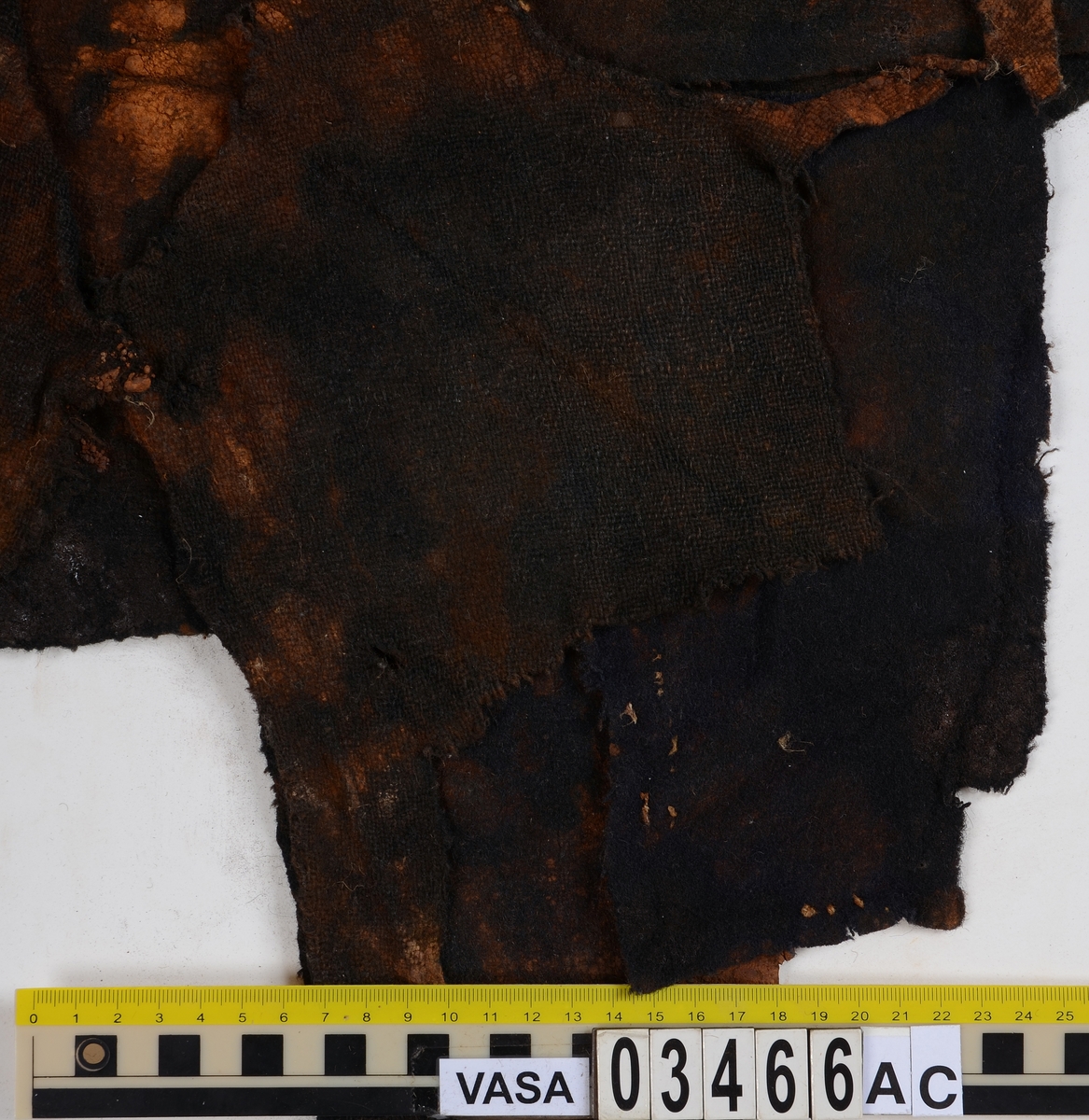 Textil. 13 tygfragment uppdelade på fyndnummer 03466a-d.
Fnr 03466a består av 10 fragment av ull vävda i tuskaft varav det största är ihopsytt av flera delar. Har varit ett par byxor med lucka fram. Originalfärgen kan ha varit blå då rester av blå färg syns på insidan av två av bitarna på de delar där sömmen varit och tyget originalyta bevarats bättre. På en av dessa bitar som tolkats som en del av en ficka på byxans vänstra sida finns rester av en tvåtrådig, s-tvinnad, z-spunnen tråd i sömmen. Byxans tyg har varit mer valkad på insidan än på utsidan. På det största fragmentet framsida syns två knapphål i linningen, och rester efter ytterligare två hål. Det kan därför konstateras att det ursprungligen har varit två knapphål på var sida på linningens mittdel där sidodelarna av linningen knäppts eller knutits fast. Vid ena knapphålet syns något som kan vara resterna av en knapp eller hyska odyl. När byxans delar läggs ihop som de en gång suttit mäter midjebredden ca 47 cm vilket ger ett midjeomfång på ca 94 cm före ihopsnörning. Av det som kvarstår av byxbenen kan konstateras att dagens (år 2015) längd på vänster byxben är 60 cm från linningens övre kant och ner. Höger byxben är i dagsläget endast 43 cm långt. 
På fnr 03466b syns rester av söm i en av kanterna. Fragmentet är av ull och vävt i 2/2-kypert. Lappen har en söm i en cirkel mitt på tyget som troligen är resterna efter den söm som sytt fast lappen runt ett hål på byxan. Kan ha varit en lagningslapp på byxans högra knä som uppvisar motsvarande söm kring ett cirkelformat hål. 
Fnr 03466c är ett fragment av ull vävt i lite finare tuskaftsväv än fnr 03466a. Fragmentet har hål efter sömmar i kanterna samt delar av en ljus tråd i en av kanterna. Blå färg syns där tyget varit skyddat där sömmen varit. Fragmentet kan ha varit en innerficka bak på byxans vänstra sida. Det finns en söm i byxans tyg där som överensstämmer med lappens söm.
Alla kanterna på fnr 03466d är invikta och har hål efter söm som om biten varit fastsydd på ett annat tygstycke. Fragmentet är av ull och vävt i samma sorts tuskaftsväv som byxan. Kan ha varit en lagningslapp på byxan över ett hål i byxtyget på den plats där vänster innerficka bak är placerad.