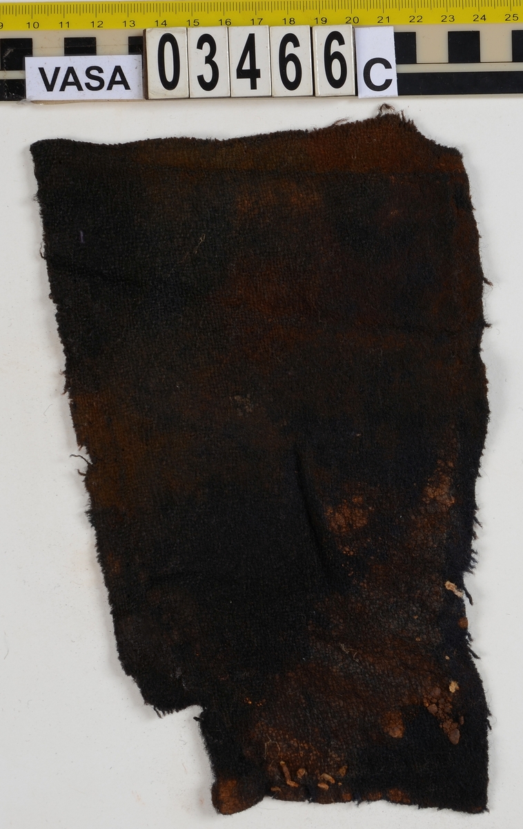 Textil. 13 tygfragment uppdelade på fyndnummer 03466a-d.
Fnr 03466a består av 10 fragment av ull vävda i tuskaft varav det största är ihopsytt av flera delar. Har varit ett par byxor med lucka fram. Originalfärgen kan ha varit blå då rester av blå färg syns på insidan av två av bitarna på de delar där sömmen varit och tyget originalyta bevarats bättre. På en av dessa bitar som tolkats som en del av en ficka på byxans vänstra sida finns rester av en tvåtrådig, s-tvinnad, z-spunnen tråd i sömmen. Byxans tyg har varit mer valkad på insidan än på utsidan. På det största fragmentet framsida syns två knapphål i linningen, och rester efter ytterligare två hål. Det kan därför konstateras att det ursprungligen har varit två knapphål på var sida på linningens mittdel där sidodelarna av linningen knäppts eller knutits fast. Vid ena knapphålet syns något som kan vara resterna av en knapp eller hyska odyl. När byxans delar läggs ihop som de en gång suttit mäter midjebredden ca 47 cm vilket ger ett midjeomfång på ca 94 cm före ihopsnörning. Av det som kvarstår av byxbenen kan konstateras att dagens (år 2015) längd på vänster byxben är 60 cm från linningens övre kant och ner. Höger byxben är i dagsläget endast 43 cm långt. 
På fnr 03466b syns rester av söm i en av kanterna. Fragmentet är av ull och vävt i 2/2-kypert. Lappen har en söm i en cirkel mitt på tyget som troligen är resterna efter den söm som sytt fast lappen runt ett hål på byxan. Kan ha varit en lagningslapp på byxans högra knä som uppvisar motsvarande söm kring ett cirkelformat hål. 
Fnr 03466c är ett fragment av ull vävt i lite finare tuskaftsväv än fnr 03466a. Fragmentet har hål efter sömmar i kanterna samt delar av en ljus tråd i en av kanterna. Blå färg syns där tyget varit skyddat där sömmen varit. Fragmentet kan ha varit en innerficka bak på byxans vänstra sida. Det finns en söm i byxans tyg där som överensstämmer med lappens söm.
Alla kanterna på fnr 03466d är invikta och har hål efter söm som om biten varit fastsydd på ett annat tygstycke. Fragmentet är av ull och vävt i samma sorts tuskaftsväv som byxan. Kan ha varit en lagningslapp på byxan över ett hål i byxtyget på den plats där vänster innerficka bak är placerad.