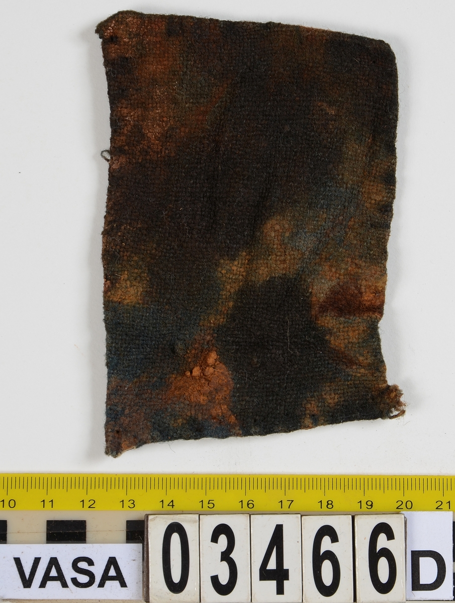 Textil. 13 tygfragment uppdelade på fyndnummer 03466a-d.
Fnr 03466a består av 10 fragment av ull vävda i tuskaft varav det största är ihopsytt av flera delar. Har varit ett par byxor med lucka fram. Originalfärgen kan ha varit blå då rester av blå färg syns på insidan av två av bitarna på de delar där sömmen varit och tyget originalyta bevarats bättre. På en av dessa bitar som tolkats som en del av en ficka på byxans vänstra sida finns rester av en tvåtrådig, s-tvinnad, z-spunnen tråd i sömmen. Byxans tyg har varit mer valkad på insidan än på utsidan. På det största fragmentet framsida syns två knapphål i linningen, och rester efter ytterligare två hål. Det kan därför konstateras att det ursprungligen har varit två knapphål på var sida på linningens mittdel där sidodelarna av linningen knäppts eller knutits fast. Vid ena knapphålet syns något som kan vara resterna av en knapp eller hyska odyl. När byxans delar läggs ihop som de en gång suttit mäter midjebredden ca 47 cm vilket ger ett midjeomfång på ca 94 cm före ihopsnörning. Av det som kvarstår av byxbenen kan konstateras att dagens (år 2015) längd på vänster byxben är 60 cm från linningens övre kant och ner. Höger byxben är i dagsläget endast 43 cm långt. 
På fnr 03466b syns rester av söm i en av kanterna. Fragmentet är av ull och vävt i 2/2-kypert. Lappen har en söm i en cirkel mitt på tyget som troligen är resterna efter den söm som sytt fast lappen runt ett hål på byxan. Kan ha varit en lagningslapp på byxans högra knä som uppvisar motsvarande söm kring ett cirkelformat hål. 
Fnr 03466c är ett fragment av ull vävt i lite finare tuskaftsväv än fnr 03466a. Fragmentet har hål efter sömmar i kanterna samt delar av en ljus tråd i en av kanterna. Blå färg syns där tyget varit skyddat där sömmen varit. Fragmentet kan ha varit en innerficka bak på byxans vänstra sida. Det finns en söm i byxans tyg där som överensstämmer med lappens söm.
Alla kanterna på fnr 03466d är invikta och har hål efter söm som om biten varit fastsydd på ett annat tygstycke. Fragmentet är av ull och vävt i samma sorts tuskaftsväv som byxan. Kan ha varit en lagningslapp på byxan över ett hål i byxtyget på den plats där vänster innerficka bak är placerad.