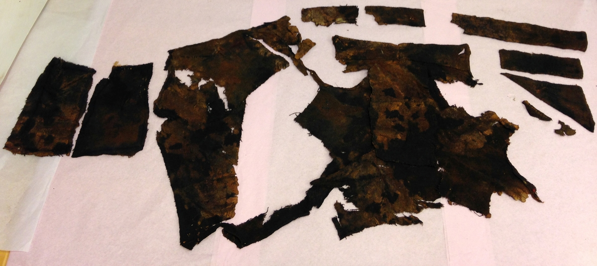 Textil. 13 tygfragment uppdelade på fyndnummer 03466a-d.
Fnr 03466a består av 10 fragment av ull vävda i tuskaft varav det största är ihopsytt av flera delar. Har varit ett par byxor med lucka fram. Originalfärgen kan ha varit blå då rester av blå färg syns på insidan av två av bitarna på de delar där sömmen varit och tyget originalyta bevarats bättre. På en av dessa bitar som tolkats som en del av en ficka på byxans vänstra sida finns rester av en tvåtrådig, s-tvinnad, z-spunnen tråd i sömmen. Byxans tyg har varit mer valkad på insidan än på utsidan. På det största fragmentet framsida syns två knapphål i linningen, och rester efter ytterligare två hål. Det kan därför konstateras att det ursprungligen har varit två knapphål på var sida på linningens mittdel där sidodelarna av linningen knäppts eller knutits fast. Vid ena knapphålet syns något som kan vara resterna av en knapp eller hyska odyl. När byxans delar läggs ihop som de en gång suttit mäter midjebredden ca 47 cm vilket ger ett midjeomfång på ca 94 cm före ihopsnörning. Av det som kvarstår av byxbenen kan konstateras att dagens (år 2015) längd på vänster byxben är 60 cm från linningens övre kant och ner. Höger byxben är i dagsläget endast 43 cm långt. 
På fnr 03466b syns rester av söm i en av kanterna. Fragmentet är av ull och vävt i 2/2-kypert. Lappen har en söm i en cirkel mitt på tyget som troligen är resterna efter den söm som sytt fast lappen runt ett hål på byxan. Kan ha varit en lagningslapp på byxans högra knä som uppvisar motsvarande söm kring ett cirkelformat hål. 
Fnr 03466c är ett fragment av ull vävt i lite finare tuskaftsväv än fnr 03466a. Fragmentet har hål efter sömmar i kanterna samt delar av en ljus tråd i en av kanterna. Blå färg syns där tyget varit skyddat där sömmen varit. Fragmentet kan ha varit en innerficka bak på byxans vänstra sida. Det finns en söm i byxans tyg där som överensstämmer med lappens söm.
Alla kanterna på fnr 03466d är invikta och har hål efter söm som om biten varit fastsydd på ett annat tygstycke. Fragmentet är av ull och vävt i samma sorts tuskaftsväv som byxan. Kan ha varit en lagningslapp på byxan över ett hål i byxtyget på den plats där vänster innerficka bak är placerad.