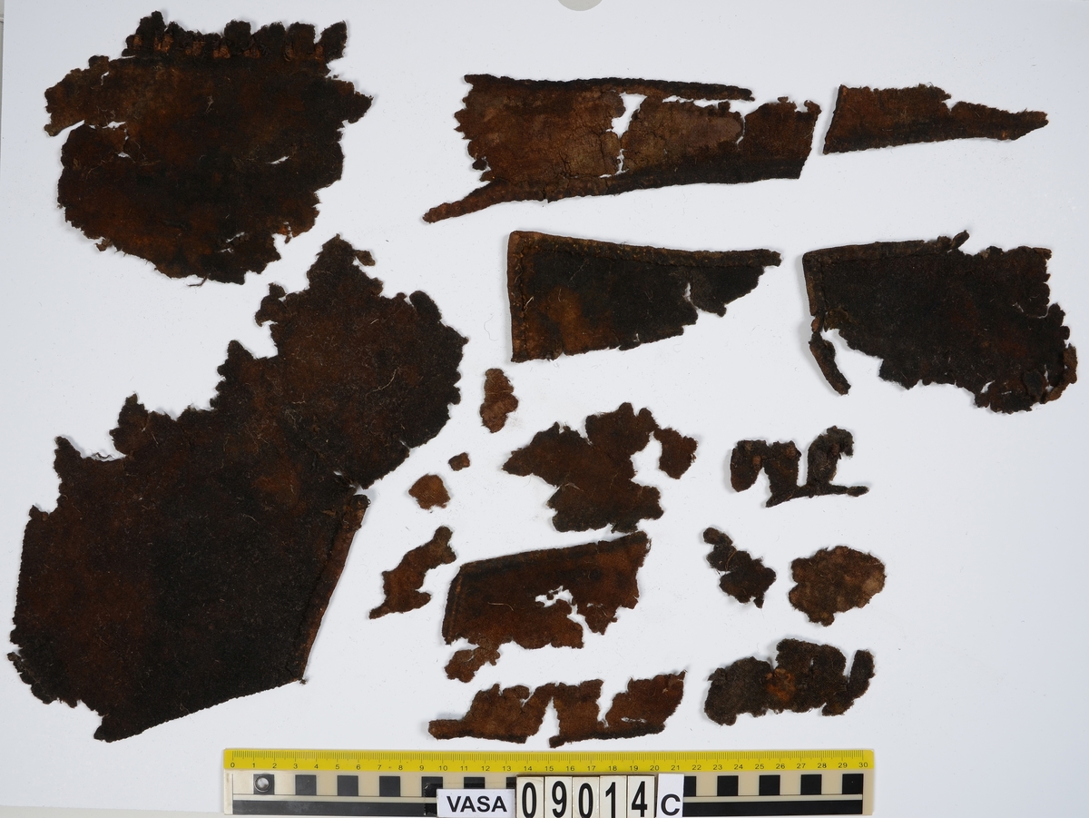 Textildokumentation 2018
Textil, fyndnumret består av mer än 50 st fragment uppdelade på 09014a-h.
Kontextgrupp ÖB20 (övriga fyndnummer i gruppen: 07732, 07762, 09228)
Relaterade fynd: Kontextgrupp ÖB17 och ÖB 18.
.
Fnr 09014a Typnr ÖB20.3:1
Sammanfattning - Ett textilfragment av ull vävt i tuskaft. Färgen är nu mörkbrun, fibrerna varierar i färg vilket tyder på att det är naturfärgad ull. Fibrerna är i vissa partier skadade på ytan och ser därför vita ut. Väven är tunn, jämn och fin och har inga spår av ytbehandling. Fragmentet har tre tillskurna kanter, två av dessa har spår av tre olika sömmar (söm 1: kvicksöm/förstygn). Formen tyder på att det är ett skört till en tröja. Andra delar: Jämför med 07726. 
.
Fnr 09014a Typnr ÖB20.4:1
Sammanfattning - Ett textilfragment av ull vävt i tuskaft. Färgen är nu mörkbrun, originalfärg okänd. I mikroskop ser tyget blåsvart ut. Väven är mycket jämn och slät. I sömsmånen på sida A finns bevarad lugg, på sida B är luggen så välbevarad att bindningen inte syns. Fragmentet har två tillskurna kanter med hål och avtryck av sömmar, även trådrester finns (söm 1: kaststygn - fälld kant, söm 2: ev förstygn - sammanfogande, söm 3: kaststygn - fälld kant). På sida B finns blåfärgad ull och silkesfibrer. 
.
Fnr 09014a Typnr ÖB20.5:1
Sammanfattning - Ett textilfragment av ull vävt i tuskaft. Väven är jämn och tät. En av kanterna är troligen tillskuren, inga sömrester finns. Tyget har inga spår av ytbehandling. På ytan finns en del bastfibrer. Relaterade fynd: Vävens struktur liknar ÖB20.4:1 men tygerna har ändå olika karaktär och detta fragment saknar lugg. 
.
Fnr 09014a Typnr ÖB20.6:1
Sammanfattning - Ett textilfragment av ull vävt i tuskaft. Färgen är nu brun, originalfärg är okänd men möjligen naturfärgad, fibrerna är mycket släta och varierar i färg från brun till nästan svart. Fragmentet är litet och mycket skadat, endast några få bindepunkter återstår vilket försvårar mätningar. 
.
Fnr 09014a Typnr ÖB20.7:1
Sammanfattning - Ett textilfragment av  ull vävt i tuskaft. Färgen är svartbrun, originalfärg okänd men möjligen naturfärgad då fibrernas färg varierar. Inga tydliga spår av ytbehandling. Väven är jämn men garnet är hårt spunnet vilket ger tyget en kypertkaraktär (falsk kypert). Fragmentet har inga tillskurna kanter eller sömmar. På ytan finns enstaka vita silkesfibrer.
.
Fnr 09014a Typnr ÖB20.8:1
Sammanfattning - Ett textilfragment av ull. Fragmentet är mycket litet och dåligt bevarat så bindningen är svår att avgöra, troligen 2/2 kypert. Färgen är svart, möjligen naturfärgad ull då fibrerna varierar något i färg. Tyget har ingen synlig ytbehandling. Garnet är hårt spunnet vilket ger tyget en speciell karaktär. 
.
Fnr 09014b Typnr ÖB17.1:8-11
Sammanfattning - Fyra textilfragment av ull vävt i tuskaft. Färgen är mörkbrun, originalfärg okänd men den bruna tonen slår mot röd. Tre av fragmenten har tillskurna kanter och spår av sömmar. Andra delar, som troligen kommer från samma tröja, är fnr 03261a och d, 09014h samt 09228a. 
Fragment ÖB17.1:9 - Fragmentet har två tillskurna kanter varav den ena har spår av en söm. Fragmentet är troligen sidostycket till en tröja. Den tillskurna kanten har då varit monterad mot tröjans bakstycke och bör ha hört samman med fyndnummer 03261d, vilket är ett bakstycke till en tröja.
Fragment ÖB17.1:10 - Fragmentet är mycket skadat men är troligen av typen ÖB17.1. Formen är densamma som för del av fragment ÖB17.1:9 och kan därför vara en dubblering av detsamma, alternativt det motstående sidostycket.
.
Fnr 09014c Typnr ÖB20.9:1-19
Sammanfattning - Nitton textilfragment av ull vävt i tuskaft. Färgen är brun, originalfärg okänd. Sida B har en kort tät lugg som döljer bindningen. På sida A finns ingen lugg och bindningen är synlig. Fjorton av fragmenten har tillskurna kanter och sömmar. Tre av fragmenten har sydda knapphål. Flera fragment är dräktdelar, till exempel finns en fragmentarisk krage. På ytan på flertalet fragment finns fibrer i flera klara färger, till exempel rosa, röd och blå. Vita fibrer finns i stor mängd, kan vara recenta.

Fragment ÖB20.9:1 - Fragmentet har flera tillskurna kanter med avtryck och hål efter olika typer av sömmar (söm 1: ev. sammanfogande, söm 2: kaststygn - fåll, söm 3: kaststygn - sammanfogande, söm 4: kaststygn - sammanfogande, söm 5: osäkert, söm 6: osäkert), trådrester finns. Kanterna och sömmarnas karaktär gör att sida A bedöms vara plaggets rätsida/utsida. Några hål som ej är stygnhål men kan vara original finns. I fragmentet sitter fast två stycken silkestrådar (söm 7: silkestråd med knut, fäst i tyget, söm 8:  silkestråd med ögla, fäst i tyget). Det är samma typ av tråd som knapphålen på fragment ÖB20.9:2 är sydda med. På sida A finns ett område med extra mycket korrosion och även en långsmal vitaktig avlagring.

Fragment ÖB20.9:2 - Fragmentet har inga bevarade tillskurna kanter, dock finns en någorlunda välbevarad rad med knapphål. Samtliga knapphål är trasiga i ena sidan så deras ursprungliga storlek går ej att avgöra. Knapphålen är sydda med knapphålsstygn i silkestråd och sitter med 13-15 mm mellanrum (c/c mått). Det tvärställda tränsen består av ca 10 stygn och syns ej på sida B, tränsens knutar sitter vända från knapphålet. På sida B anas rester av bastfiber under knapphålsstygnen, troligen från en dubblering av tyget eller ett foder. 

Fragment ÖB20.9:3 - Fragmentet har fyra stycken knapphål, varav ett är helt. Knapphålen är skurna på skrådden. Eventuellt finns spår av en söm längs en del av kanten. Knapphålsstygn och trådrester är mycket fragmentariska.

Fragment ÖB20.9:4 - Fragmentet har tre stycken knapphål.  Knapphålen är sydda med knapphålsstygn i silkestråd. Knapphålens längd är 19-20 mm och de sitter med 18-22 mm mellanrum (c/c mått). Eventuellt är fragmentets ena kant en sömsmån, kan ha varit monterad mellan andra tyglager (kanten har avvikande färg). Under knapphålsstygnen på sida B finns rester av annat material, eventuellt bastfiber. Bastfibrer finns också utspridda över ytan på sida B, kan vara rester av en dubblering till tyget eller ett foder, se fragment 9:2. På sida A finns avlagringar och korrosion. 

Fragment ÖB20.9:5 - Fragmentet har bristningar i tyget som ser ut som knapphål, dock finns inga rester av stygnhål eller tråd. Fragmentets ena kant är troligen en sömsmån, kan ha varit monterad mellan andra tyglager (kanten har avvikande färg). Enstaka spår av söm och tråd finns längs kanten (söm 1), dessa är mycket otydliga. På fragmentets andra (mycket trasiga) kant finns ett mycket tydligt stygnhål. Fragment 9:4 och 9:5 ser ut att kunna höra ihop, dock går trådsystemen åt olika håll så det är inte fallet.

Fragment 9:6-9 - Till det stora fragmentet 9:6 hör det lilla lösa fragmentet 9:7. I fragment 9:6 sitter två fragment fastsydda, 9:8 och 9:9. De sammansydda fragmenten har tre tillskurna kanter och spår av minst fem sömmar (söm 1: kaststygn, söm 2: förstygn el. liknande - sammanfogande, söm 3: kaststygn, söm 4: sammanfogande, fäster ihop 9:6 med 9:8 och 9:9, söm 5: två stygnhål), trådrester finns. Det är troligt att 9:8 och 9:9 är delar av samma tillskurna bit, och att den utgjort en dubblering till 9:6 (inklusive 9:7). Den tillskurna formen ser ut att vara en krage. Fragmentet skulle då vara en halv krage, bevarad från framkant till nästan mitt bak. På sida A finns en beläggning på delar av ytan. På sida B finns en mängd fibrer i klara färger: blå, vit, rosa, tegelröd, orange, karminröd, grön, mörkgul och grå. fibrerna ligger ytligt med nästan samtliga sitter delvis fast i tygets lugg eller har en eller båda ändar nere i vävens bindning. Fibrerna bör därför vara ursprungliga. Sida A har en mindre mängd av nämnda fibrer, mest förekommande på ytan av fragment 9:8 och 9:9.

Fragment ÖB20.9:10 - Fragmentet har två tillskurna kanter. Dessa är invikta mot sida B som en 6-7 mm bred fåll som är fastsydd med kaststygn, trådrester finns (söm 1 och 2). Fragmentet kan vara ett skört till en tröja. På båda sidors yta finns fibrer i klara färger: grå, ljusblå, orange, orangebrun, vitgul, blå svart, vit, röd, korallröd och rosa.

Fragment ÖB20.9:11 - Fragmentet har tre tillskurna kanter med sömmar. Två av kanterna är invikta och fastsydda mot sida B, som fållar (söm1: kaststygn - fåll, söm 2: kaststygn - fåll). Stygnen går ej igenom till sida A men sömmen syns som bucklor i fragmentets yta. Söm 3 längs den tredje kanten har troligen varit en sammanfogande söm sydd med kaststygn. Stygn och trådrester finns bevarade från samtliga sömmar. Under fållen vid söm 1 finns ett litet fragment av en annan textiltyp. Troligen ursprungligen en dubblering eller liknande. Fragmentet är ca 10x5 mm stort och har fått typnummer ÖB20.16:1. På ytan på sida B finns enstaka bastfibrer.

Fragment ÖB20.9:12 - Fragmentet har två tillskurna kanter med sömmar (söm 1: ev kaststygn, söm 2: osäker). Vid söm 2 är sömsmånen vikt mot sida B. Vid söm 1 är kanten inte vikt. Fragmentet har samma form som ÖB20.9:6, "kragen".

Fragment ÖB20.9:13 - Fragmentet har två tillskurna kanter med sömmar (söm1: sammanfogande, söm 2: osäkert). De två övriga kanterna är eventuellt också de tillskurna men spår av eventuella sömmar är för otydliga. Enstaka fibrer med klara färger (rosa, blå, vit) finns på båda sidor, fler på sida B. Vid söm 2 finns på ett ställe en fiberknippa som kommer upp ur vävens bindning, kan vara rester av en tråd och är eventuellt halsplat silke. 

Fragment ÖB20.9:14 - Fragmentet har en tillskuren kant med söm (söm1: ev sammanfogande). I stygnhålen finns rester av tråd.
.
Fnr 09014c Typnr ÖB20.10:1
Sammanfattning - Ett textilfragment av ull vävt i tuskaft. Färgen är brun, originalfärg okänd. Sida B har en kort och tät lugg, bindningen syns bara på sida A. Längs ena kanten finns tre stycken tydliga hål. Hålen är gjorda med ett verktyg som skär av fibrerna snarare än tränger undan dem.
.
Fnr 09014c Typnr ÖB20.16:1
Sammanfattning - Fragmentet är mycket litet och sitter inuti den invikta fållen på fragment 09014c ÖB20.9:11, det sticker bara fram någon millimeter. Fragmentet är mycket skört. Bevarandegrad och storlek gör mätningar svåra. Det är olika garn i trs 1 och trs 2. Sida B är täckt av en lugg, på sida A syns bindningen, om än fragmentariskt.
.
Fnr 09014d Typnr ÖB18.16:2-8
Ej dokumenterad
.
Fnr 09014d Typnr ÖB20.11:1-2
Ej dokumenterad
.
Fnr 09014e Typnr ÖB20.12:1-4
Sammanfattning - Fyra textilfragment av ull vävt i tuskaft. Färgen är brun med inslag av täckhårsfibrer i en mörkare brun färg, originalfärg okänd. Fragmenten är mycket fragmentariska med trs 1 bättre bevarat än trs 2. Enbart på en del av fragment ÖB20.12:1 finns trs 2 samt bindningen bevarad. Utspridda över tygets yta finns grova bastfibrer samt korrosionsfläckar. 
.
Fnr 09014f Typnr ÖB20.13:1
Sammanfattning - Ett textilfragment av ull vävt tuskaft. Färgen är mörkbrun, fibrerna varierar mellan ljus och mörk brun. Originalfärg okänd, möjligen naturfärgad ull. Garnet är hårt spunnet vilket ger väven en särskild karaktär, sida A har en mer strukturerad ytan än sida B. Fragmentet har inga tillskurna kanter. På sida A finns enstaka röda och gulgröna silkesfibrer, samt bastfibrer. På sida B finns enstaka blå, vita och rosa silkesfibrer.
.
Fnr 09014g Typnr ÖB20.14:1-3
Sammanfattning - Tre textilfragment av ull och bastfiber vävt i tuskaft. Färgen är beige för bastfibern (trs 1) och svartblå för ullen (trs 2). Trs 2 är mycker grövre än trs 1. Trs 1 är nästan helt försvunnet. Två av fragmenten har tillskurna kanter och ett har spår av en sömsmån. På ytan av fragmenten finns en stor mängd bastfiber samt en vit/rosa tuss med silke. På fragment 14:2 finns några intensivt blå ullfibrer.
.
Fnr 09014h Typnr ÖB17.1:12-13
Sammanfattning - Två textilfragment av ull vävt i tuskaft. Färgen är brun, originalfärg okänd. Det större fragmentet är tillskuret och har spår av sömmar. Fragmentet är en ensömsärm med sömmen placerad mitt bak på ärmen. På ytan finns stora rostfläckar. Andra delar: Hör troligen (trots färgskillnad) till skelettet Beatas tröja, se fnr 03261a-d, 09228a, 09014b och h.