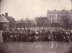 Elementarskolan i Trelleborg, firandet av Gustaf Adolfsfesten 1894, Södra skolgården med rektor, lärare och elever, foto fr. N mot S. den (6nov 1894?) i fonden hamnen. Skolgården begränsas av ett träplank, med portgrind. Till höger Rektor Sjövalls hönshus vid planket, därintill ett boketräd(Fagus) Husen: från vänster 1: Skolans hemlighus (under skiffertak) sammanbyggt med 2: Rektor Sjövalls tvättstuga, m duvslag på taket. 3: Skräddare Ant. Nilssons hus 1-vån. med 1st. skorsten, bel. Å tomt nr. 19 (59B) inom kv. Herkules. 4: Tryckarehusen 2 våningsbyggn. Uppf. Av boktryckarna Sandberg och Jönsson å tomt nr 20 (61B) inom kv. Herkules. Äldre personer från vänster: 1: Rektor Hjalmar Sjövall, fil dr 2: Kollega Elof ,fil dr 3: Kollega John Smith, fil dr 4: Kollega Alexander Rydberg, pastor
5: Flickskolelärarinnan fröken Charlotte Jönsson i spetssammetshatt (nackhatt)    6:Lärarinnan fröken Mimmi Rosqvist i bred hatt med sidenband 7: Lärarinnan Emmy Ohklander dotter av Framför det förstnämnda huset, synas den med halm vinterinklädda träpumpen
5146 Axel Andersson.