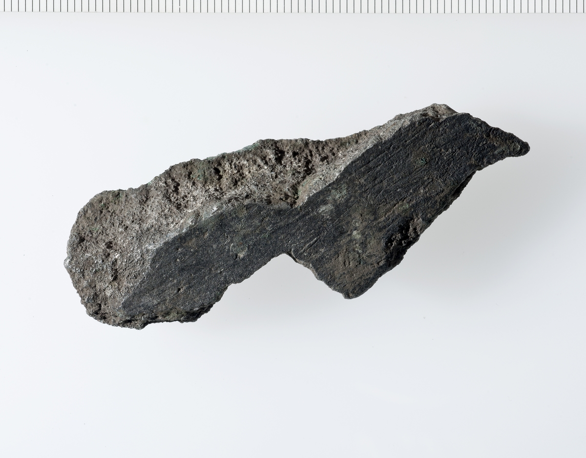 Svakt buet avlangt fragment av støpt materiale. Fragmentets buede sider er glatte, bruddsidene er porøse.  Fragmentet har for tykke vegger til å være fra en gryte, og gir en for stor diameter til å kunne være fra noen kanon. Derfor antatt fragment fra klokke. 