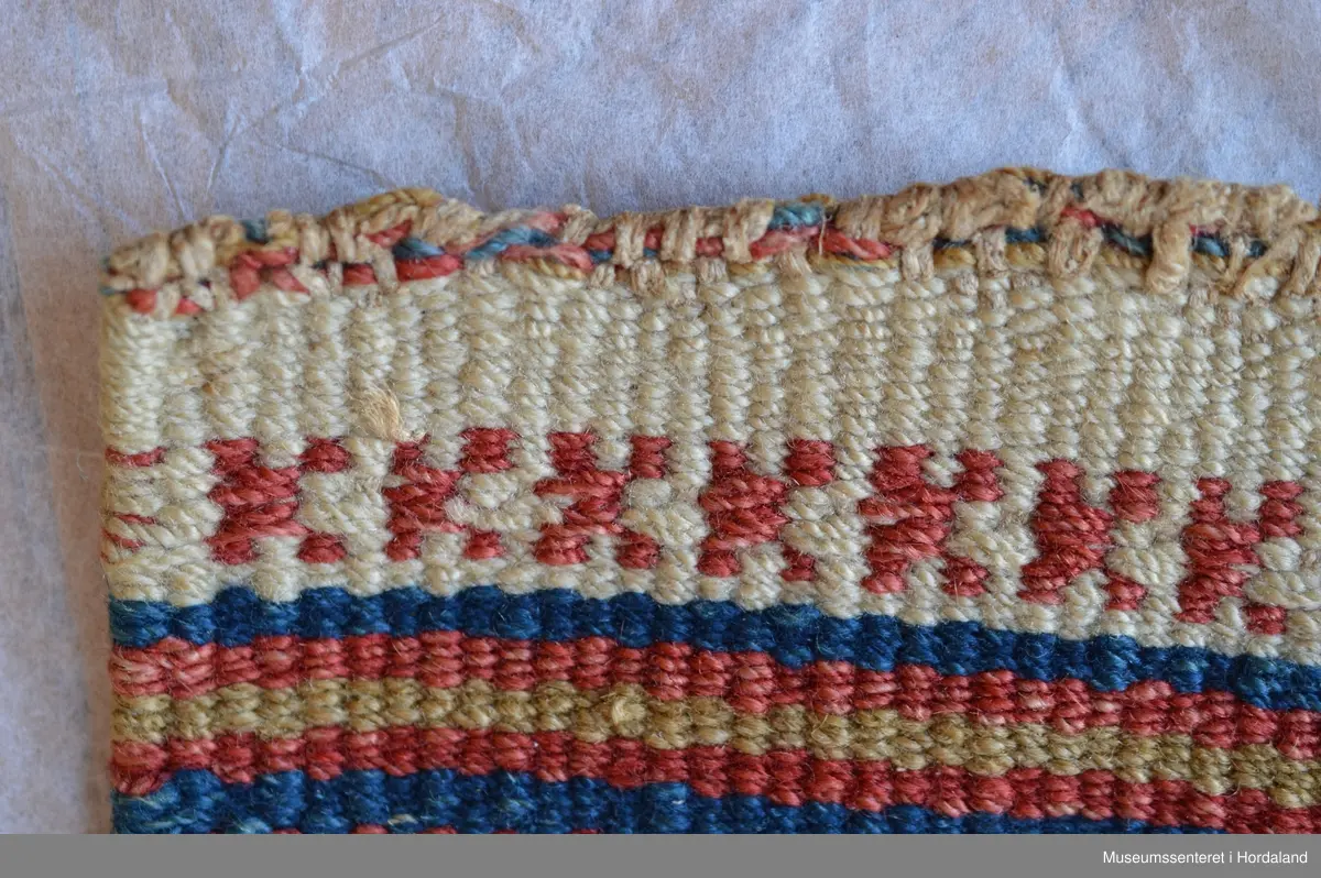 Åkle som er samansett av 8 border i halvsmett og enklare border innimellom.Bukta på renningen ser ein oppe på teppet. Dei første innslaga er delivis bortsletne. Nede på teppet er det kasta over for hand; 1-2 cm lange frynser.