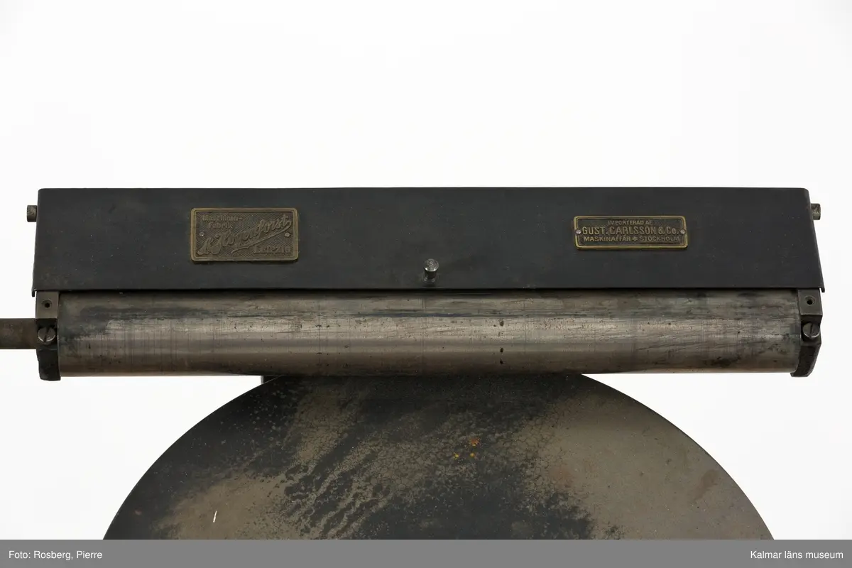KLM 45377:60. Tryckpress, Trä och metall. Digelpress av svartmålad metall, stommen av gjutjärn. På digelpressens övre del sitter två metallplåtar. På den ena plåten står det Maschinenfabrik A Hogenforst Leipzig. På den andra står det Importerad av Gust. Carlsson&Co Maskinaffär Stockholm. Digelpressen är monterad på ett litet träbord som förmodligen är byggt för pressen. Det är delvis målat i vitt.