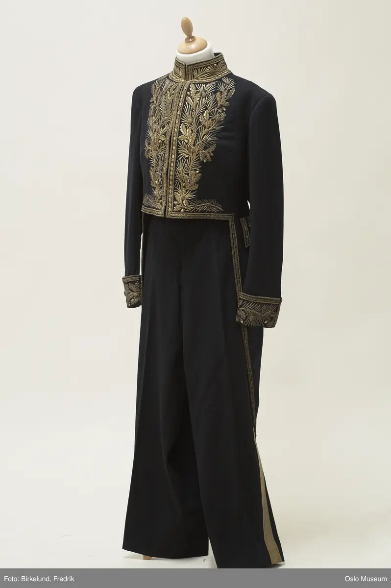 Gallauniform til ambassadør.
A: Jakke-kjole. Jakken er ettersittende foran m. høy krage som lukkes m. hekter. Lang snipp bak. Dekorbånd langs sidene og langs snipp. Dekorerte lommeklaffer og dekor på midten bak. Ermer m. dekorerte mansjettoppslag. Hemper til medaljer. Gullgalloner på buksen.Dekormotiv: Rike blomsterranker m. ekeblad. Lengde foran: 41 cm.
Lengde bak: 114 cm.
B. Bukse m. nesten rette ben. Stikklommer på siden som er skjult under gallongene. Gylf m. knepping. Liten splitt i linning bak. Knapper til seler, doble foran, enkle bak. Dekorert m. gallongbånd i damaskvev. Motiv: Ekebladranker. Lengde:115 cm.
C.Dobbelsnutehatt til ambassadøruniform m. strutsefjær og gullbånd.Stoffet på brem er trukket over et papplikninde materiale. Bremmen er formet opp på sidene slik at hatten former snuter foran og bak. En "Flaggrose" er applikert på høyre side. Tvunnet gullbånd og knapp er festet over. Gulldekor på hver snute. Strutsefjær montert langs hattebrem. Gullbånd m. ekebladmotiv langs hattebrem. Høyde: 13 cm. Lengde: 46 cm. Materiale: Fløyel og papp.God tilstand. Skjør strutsefjær.
D. Hatteeske til dobbelsnutehatt i lakkert lerretstrie/seilduk. Sort. Lukkes m. lærrem på toppen. Esken har hattens form. Vattert, gult bommulsfòr inni.
