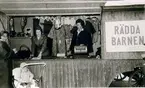 Från Rädda Barnens försäljning på Stora torget 1963. Vagnen är lånad av Lottakåren i Falköping. Rädda Barnen: Ruth Blom, född Eriksson, ordförande i Rädda Barnen, Anna Lisa Johansson, född Johnsson (i mitten), Rut Rudberg, född Svensson (längst till höger), Eva-Marie Mohlin (i barnvagnen).