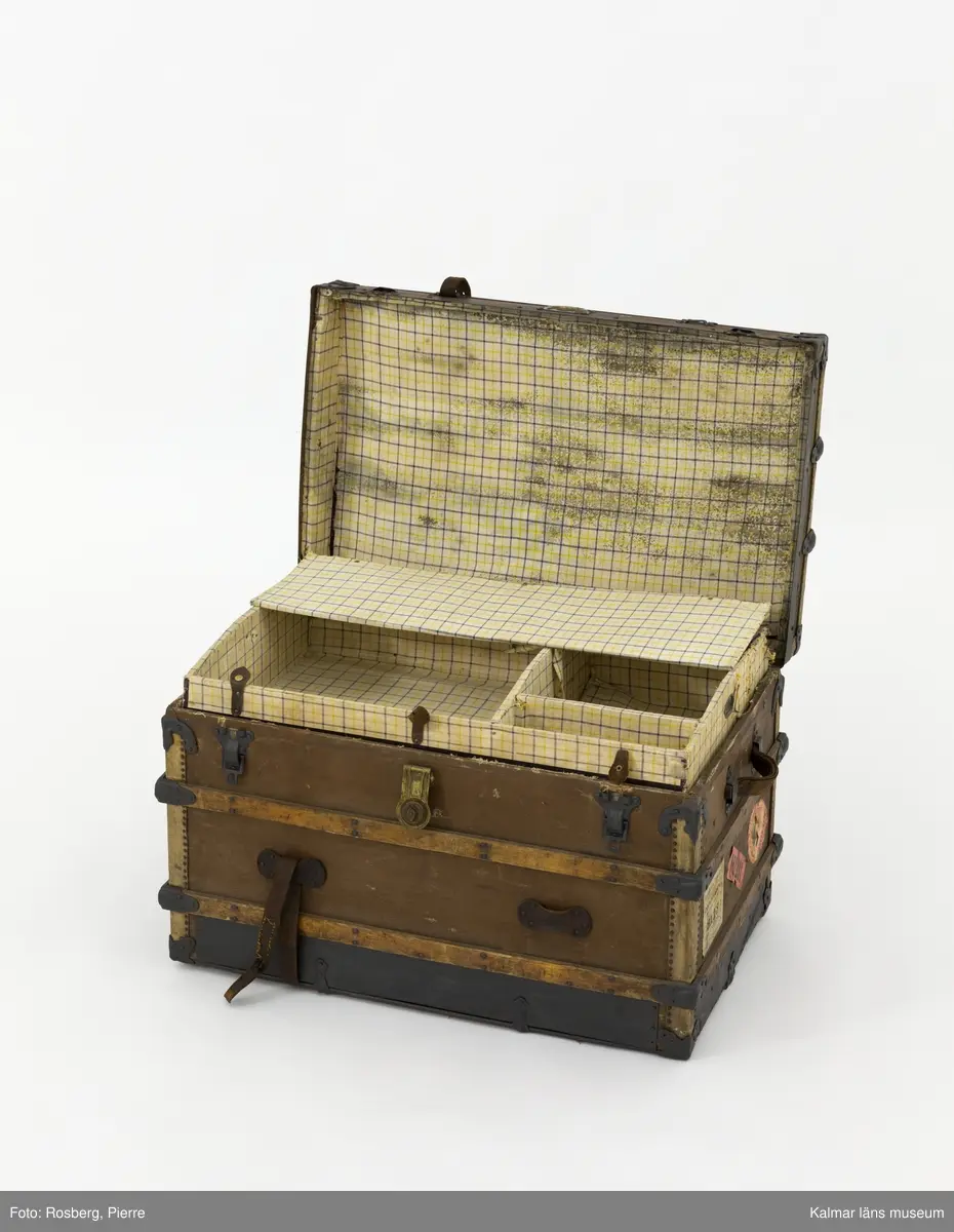 KLM 28581. Koffert, amerikakoffert, trä, väv, metall, läder. Amerikakoffert av trästomme klädd med väv. Trärribbor som är sammanfogade med plåtbeslag finns på alla sidor. Kistan hålls stängd med två bygelbeslag i metallplåt samt ett lås av mässing. Runt kistan har det funnits två läderremmar, endast en av dem finns kvar. På kistans sidor finns bärhandtag av läder. Invändigt är kistan klädd med ett vitt tyg med ränder i gult och blått som bildar rutmönster. En lös insats med tre mindre fack finns i kistans övre del. Den är av trä och klädd med tyg. Överst i kistan finns en löst lock av kartong klädd med samma tyg som resten av kistans insida.