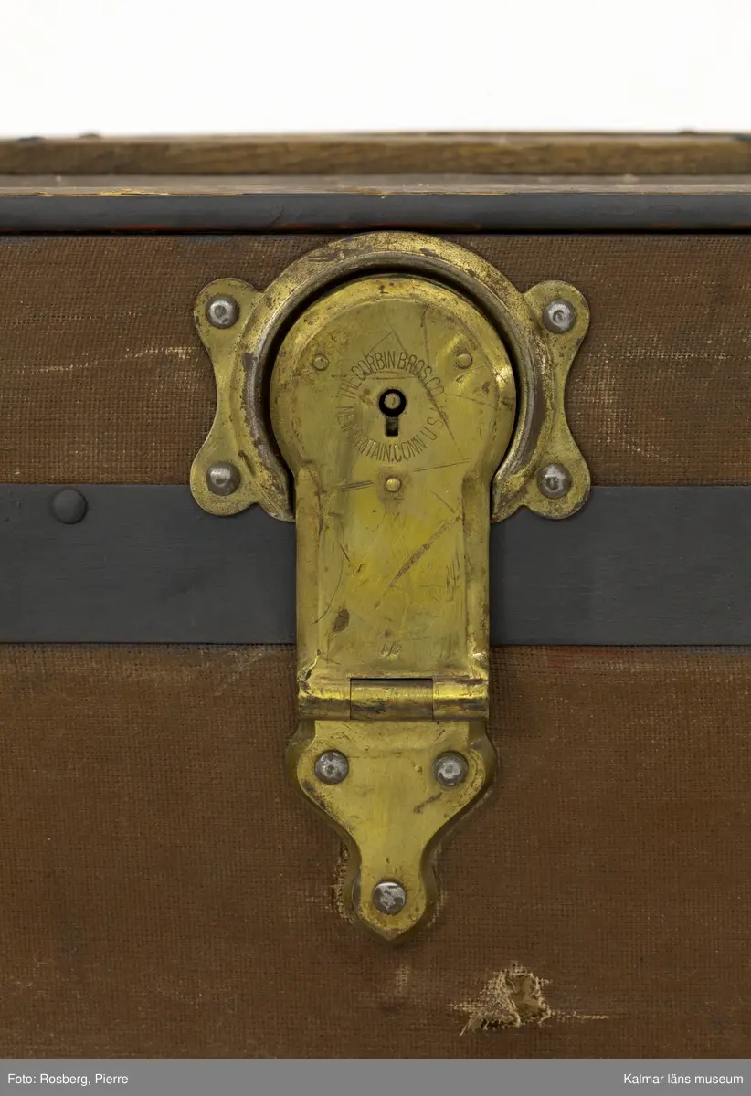 KLM 28581. Koffert, amerikakoffert, trä, väv, metall, läder. Amerikakoffert av trästomme klädd med väv. Trärribbor som är sammanfogade med plåtbeslag finns på alla sidor. Kistan hålls stängd med två bygelbeslag i metallplåt samt ett lås av mässing. Runt kistan har det funnits två läderremmar, endast en av dem finns kvar. På kistans sidor finns bärhandtag av läder. Invändigt är kistan klädd med ett vitt tyg med ränder i gult och blått som bildar rutmönster. En lös insats med tre mindre fack finns i kistans övre del. Den är av trä och klädd med tyg. Överst i kistan finns en löst lock av kartong klädd med samma tyg som resten av kistans insida.