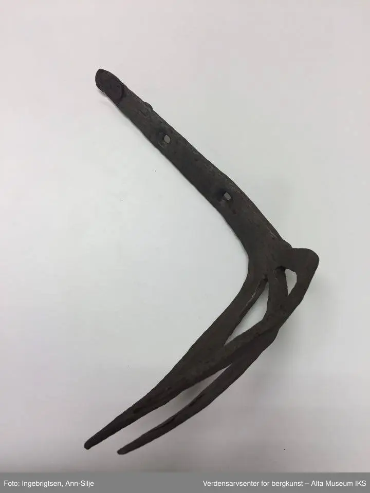 Trearmet jerngaffel. Kort skaft i samme metall. Liten krok på baksiden øverst på skaftet. To hull i skaftet (3 cm mellomtom). Hver av "armene" er ca. 20 cm lange. Skaftet er 23 cm langt.