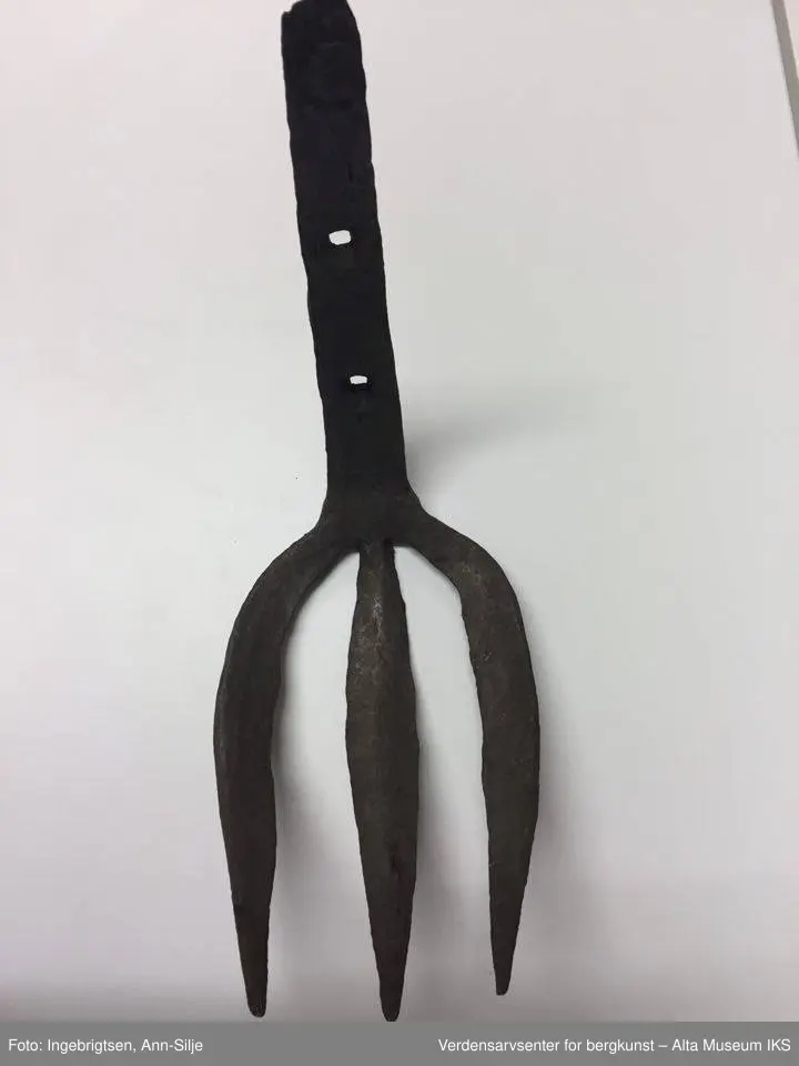 Trearmet jerngaffel. Kort skaft i samme metall. Liten krok på baksiden øverst på skaftet. To hull i skaftet (3 cm mellomtom). Hver av "armene" er ca. 20 cm lange. Skaftet er 23 cm langt.