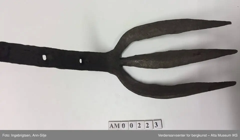 Trearmet jerngaffel. Kort skaft i samme metall. Liten krok på baksiden øverst på skaftet. To hull i skaftet (3 cm mellomtom). Hver av "armene" er ca. 20 cm lange. Skaftet er 23 cm langt.