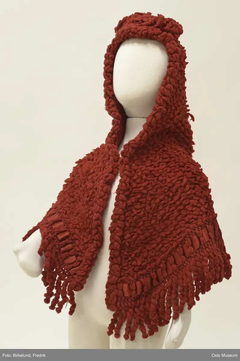 Trekantet; på toppen formet som liten kyse, nederst i ryggen oppbuet parti. Nettverk av rødt ullgarn itredd utvunnete biter som danner en tett overflate. Kysen og nedkanten m/frynser. På kysen sløyfe og nederst itredd bånd av silke. På begge sider av båndet itredd rødt silkegarn.