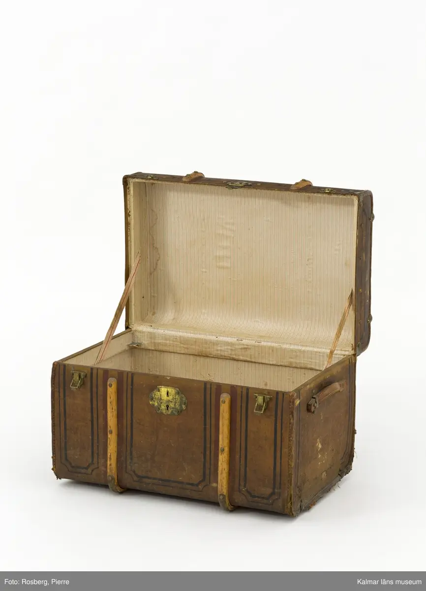 KLM 31996. Koffert, reskoffert, trä, metall, textil, läder, färg, Brunmålad, rektangulär koffert med svarta mönster och ett handtag av läder på varje gavel. På fronten ett nyckellås samt två låsspännen av mässing. Ribbor av trä med mässingsbeslag runt kofferten som skydd/stötdämpning. Inuti klädd med grön- och vitrandig papp med väv på baksidan.