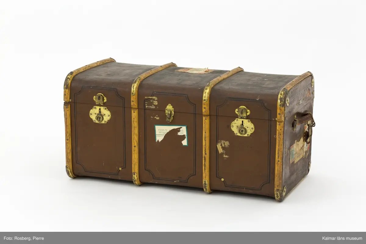 KLM 32819:4. Koffert, reskoffert, trä, metall, läder, textil. Brunmålad reskoffert av trä, belagd med väv, med svartmålad dekor. Beslag av trä och metall för stötdämpning/skydd. På varje gavel ett handtag av läder. På fronten tre låsspännen av mässing. Invändigt klädd i grönt tyg med två uttagbara förvaringskorgar av trä med spännen. I den ena är botten lös. På lockets insida klisterlapp: Senaste nyheter HANDSKAR PARAPLYER RESEFFEKTER SVEN NÄRSTAD KALMAR. 
Ovanpå locket etikett: PAKET-eller? Från Fru Hertha Jeansson, Kalmar, Kalmar C Efterkrav Kr..... Till Fru Märtha Frick Svedbergshuset Wennerbergsgatan 6 Bestämmelsestation STOCKHOLM (enligt fraktsedeln) 1 kolli.
Målat på lockets ovansida: M.J 3974 1.
På höger gavel tillverkarmärke: AKTIEBOLAGET JOHN A. GAHM GÖTEBORG,9.