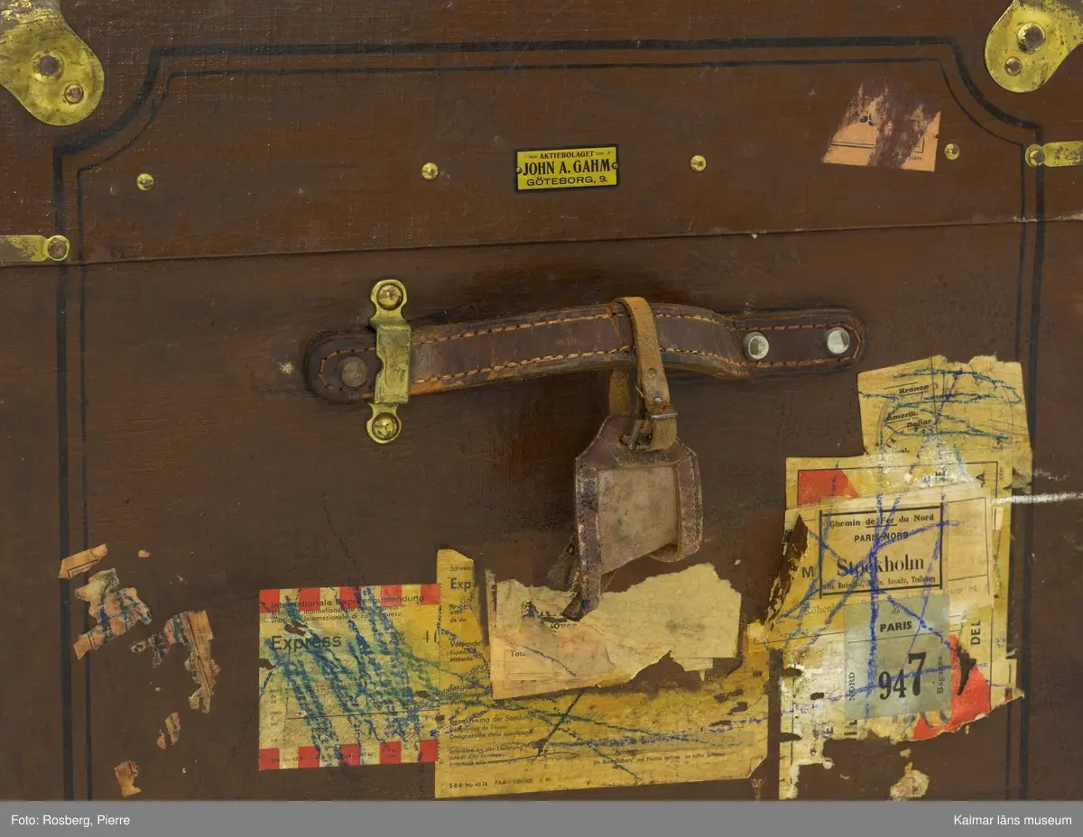 KLM 32819:4. Koffert, reskoffert, trä, metall, läder, textil. Brunmålad reskoffert av trä, belagd med väv, med svartmålad dekor. Beslag av trä och metall för stötdämpning/skydd. På varje gavel ett handtag av läder. På fronten tre låsspännen av mässing. Invändigt klädd i grönt tyg med två uttagbara förvaringskorgar av trä med spännen. I den ena är botten lös. På lockets insida klisterlapp: Senaste nyheter HANDSKAR PARAPLYER RESEFFEKTER SVEN NÄRSTAD KALMAR. 
Ovanpå locket etikett: PAKET-eller? Från Fru Hertha Jeansson, Kalmar, Kalmar C Efterkrav Kr..... Till Fru Märtha Frick Svedbergshuset Wennerbergsgatan 6 Bestämmelsestation STOCKHOLM (enligt fraktsedeln) 1 kolli.
Målat på lockets ovansida: M.J 3974 1.
På höger gavel tillverkarmärke: AKTIEBOLAGET JOHN A. GAHM GÖTEBORG,9.