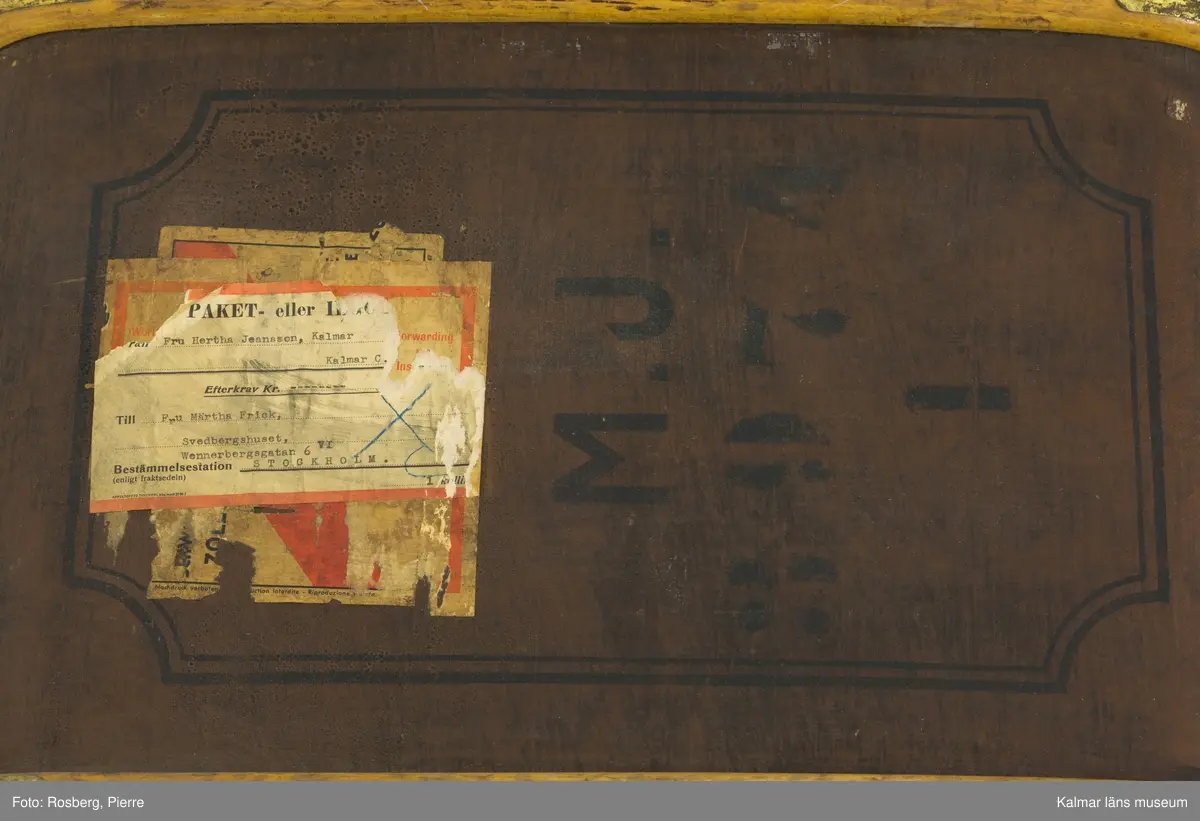 KLM 32819:4. Koffert, reskoffert, trä, metall, läder, textil. Brunmålad reskoffert av trä, belagd med väv, med svartmålad dekor. Beslag av trä och metall för stötdämpning/skydd. På varje gavel ett handtag av läder. På fronten tre låsspännen av mässing. Invändigt klädd i grönt tyg med två uttagbara förvaringskorgar av trä med spännen. I den ena är botten lös. På lockets insida klisterlapp: Senaste nyheter HANDSKAR PARAPLYER RESEFFEKTER SVEN NÄRSTAD KALMAR. 
Ovanpå locket etikett: PAKET-eller? Från Fru Hertha Jeansson, Kalmar, Kalmar C Efterkrav Kr..... Till Fru Märtha Frick Svedbergshuset Wennerbergsgatan 6 Bestämmelsestation STOCKHOLM (enligt fraktsedeln) 1 kolli.
Målat på lockets ovansida: M.J 3974 1.
På höger gavel tillverkarmärke: AKTIEBOLAGET JOHN A. GAHM GÖTEBORG,9.
