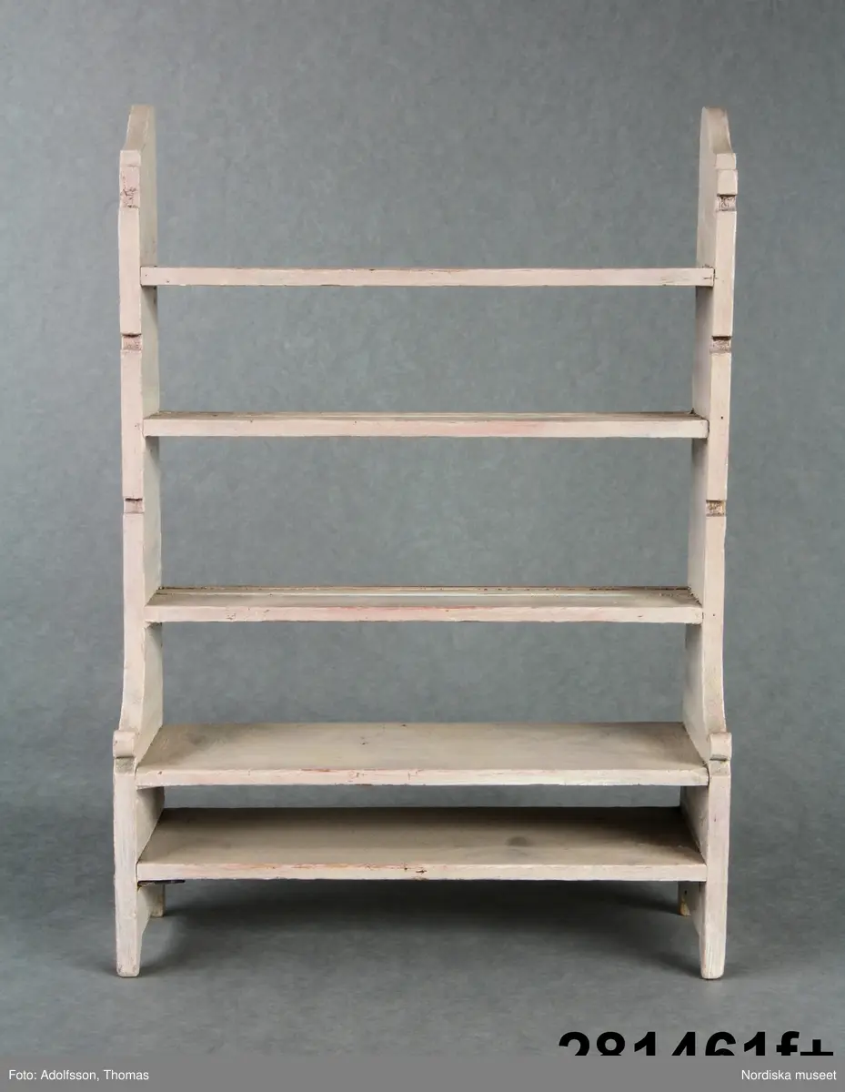 Inventering Sesam 1996-1999:
a+) Dockskåp L 104 cm, B 30 cm, H 178 cm.
Dockskåp med inventarier
 a+) Dockskåp av trä, rakt krön, korta och nedåt avsmalnande ben. Grönmålat med röda lister, dragknoppar mm. Ovanpå brunbetsat (trol. originalfärg). Underdel med tre lådvarv, överdel med glasdörrar med metallhasp upptill. Överdelen inredd till dockskåp i tre våningar med sammanlagt 7 rum. Alla rum tapetserade med olika tapeter och blindfönster av svart och vit papper klistrat på tapeterna. Nedre våning med brunrutig vaxduk som golvbeläggning. 
Dockskåpet är troligen ett ombyggt och ommålat äldre bokskåp. 
Bilaga.  
Har tillhört prinsessan Ingrid (född 1910, dotter till Gustav VI Adolf), sedemera Drottning av Danmark.