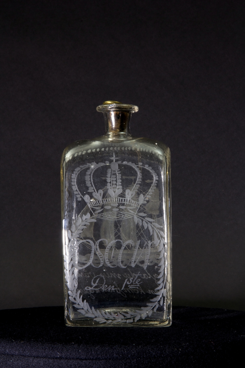 Längs flaskans övre kant en dekorativ bård av prickar. Gravyr på ena långsidan föreställande ett tremastat skepp med fulla segel. Gravyr på andra långsidan föreställande en sluten krona ovanför en lagerkrans som omsluter texten "Oscar Den 1ste". Blomstergirlanger på flaskans kortsidor.