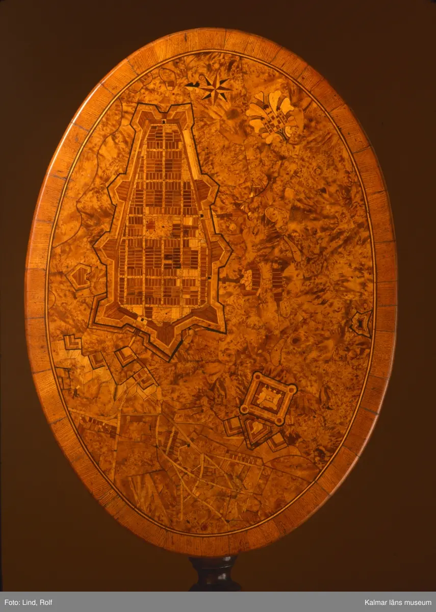 KLM 10594. Bord, fällbord. Av trä. Oval skiva av alrot med rika inläggningar, återgivande stadsplan över Kalmar stad med  Kvarnholmen och Kalmar slott, Gamla stadens gatunät, Grimskär skans, inläggning med norrpil samt Stadsporten som ett dekorativt element med blad. Pelare, svarvad och profilerad med tredelad fot. Fällbar bordsskiva med mekanism som fungerar. Datering, gustaviansk, 1700-talet slut.