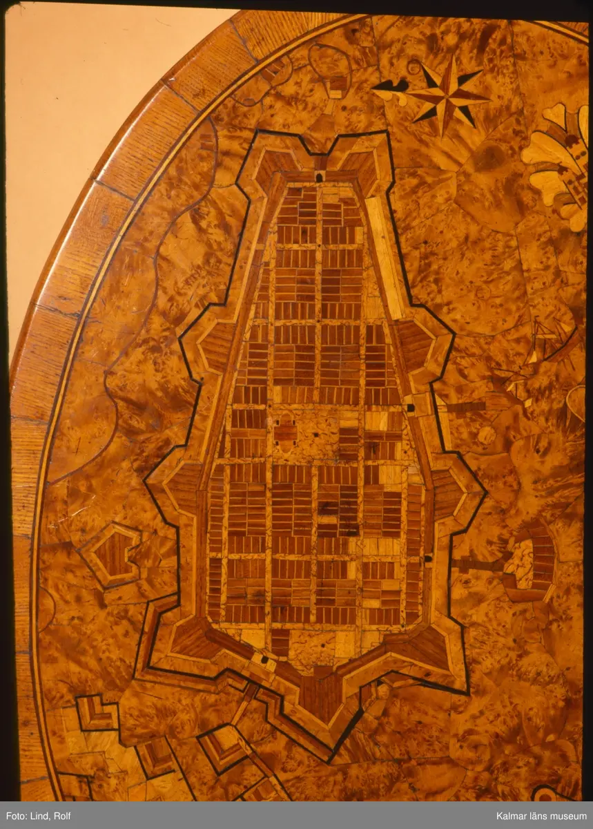 KLM 10594. Bord, fällbord. Av trä. Oval skiva av alrot med rika inläggningar, återgivande stadsplan över Kalmar stad med  Kvarnholmen och Kalmar slott, Gamla stadens gatunät, Grimskär skans, inläggning med norrpil samt Stadsporten som ett dekorativt element med blad. Pelare, svarvad och profilerad med tredelad fot. Fällbar bordsskiva med mekanism som fungerar. Datering, gustaviansk, 1700-talet slut.