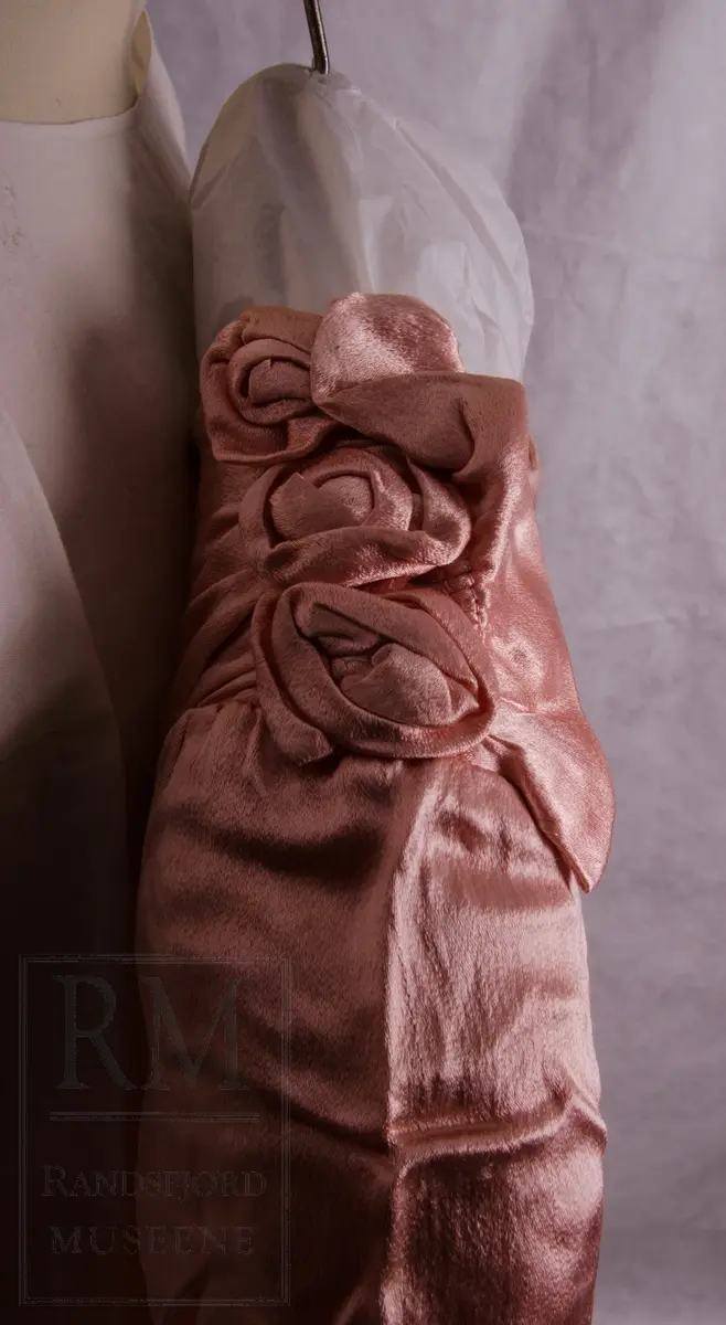 Brudekjole m/slør. 
a) Lyerød, lakserød kunstsilkesatin med creppet orange. Skjøret av 4 skrådde bredder + innfelt rynket parti bak. Søm i livet. Trykknapper i siden. Drapert knyttebelte. Enkel overdel med lukning på venstre skulder med trykknapper pyntet med 3 stoffroser og rynkekappe. Lange ermer med stikk nederst. 
b) Slør av rutevevd tyll. Hvitt med maskinbrodert blomstermotiv i hjørnene.