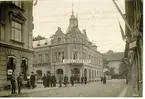 Från fältmanövern 1912. Korsningen vid Jouvins hotell (sedermera under källarmästare Bergmans regi,kallat Stadshotellet) av Järnvägsgatan /Ranta-vägen (numera S:t Olofsgatan) och  Storgatan, under manövern 1912. Paret t.v. är marinintendent Collberg med fru, dotter till provinsialläkaren S.Collin i Falköping. Den i mitten av de tre under 1:a E-et i skylten Högkvarteret är arb. Flood. Mannen bakom den lilla pojken till höger är gatuförman  Helmér.