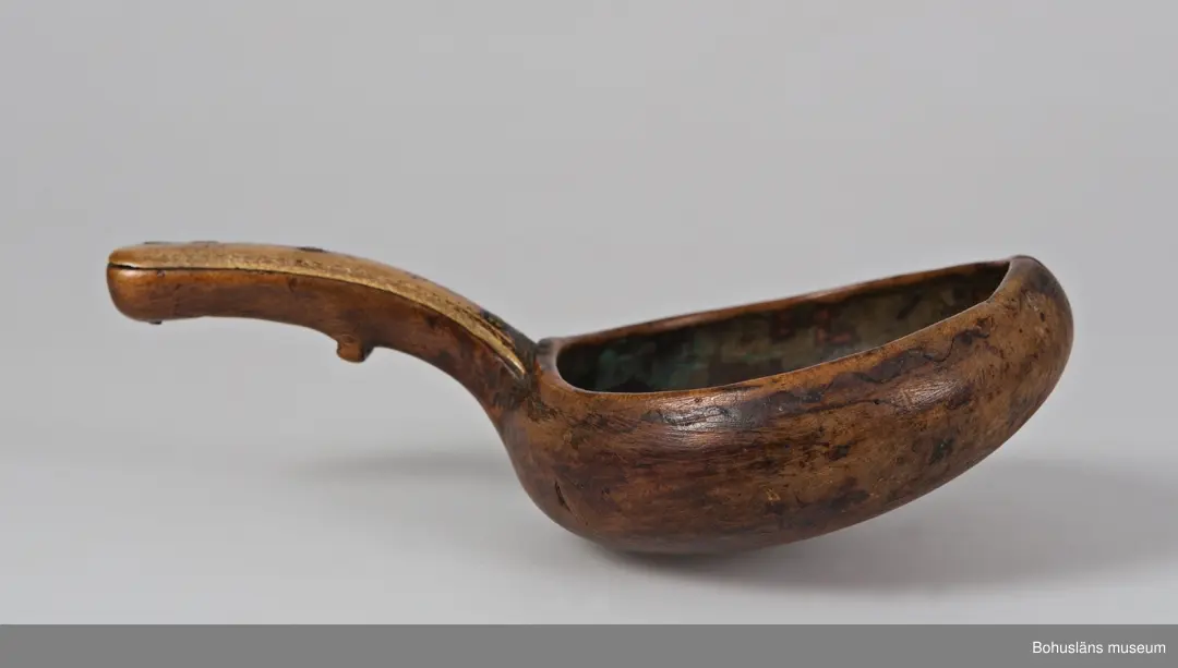 Rund skålformad kåsa med handtag skuret ur ett stycke. Mynningskanten är något indragen och mittemot handtaget är kanten upphöjd.
På utsidan kanten löper en ristad vågformad dekor.
Skaftet är böjt och bredare baktill. Ovansidan har en hornbit fastsatt med mässingsnubb. Hornbiten har ristad dekor med vågformad kant samt ett litet kors och en större korsliknande dekor.
På handtagets undersida finns ett tvärgående, utstickande parti troligtvis för att kunna hålla den lättare eller för att vila den på ett kärls kant.
Upptill på handtagets undersida finns ett inristat bomärke.
En bit av hornbiten är mörkare och sekundärt ilagad.
Kåsan har en spricka i kantens upphöjning. Inuti är det ärggröna fläckar.
Tre små hål, troligtvis efter häftstift.

Uppgifter i samband med besök från Ájtte, Svenskt Fjäll- och Samemuseum, Jokkmokk:
Lulesamisk, låg modell. Ägarnas bomärke

Ur handskrivna katalogen 1957-1958:
Träskål
Diam.: 8,5. Skopa el. Slev av trä. Föremålet helt. Lappland

Lappkatalog: 7