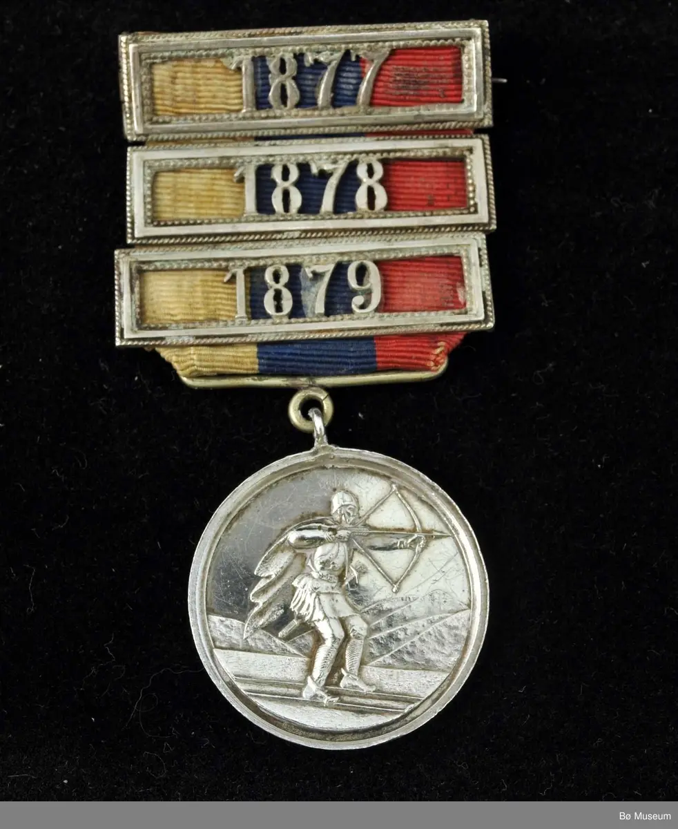 Rund medalje med bånd og nål. Skiløper med pil og spent bue på den ene siden, på den andre tekst.