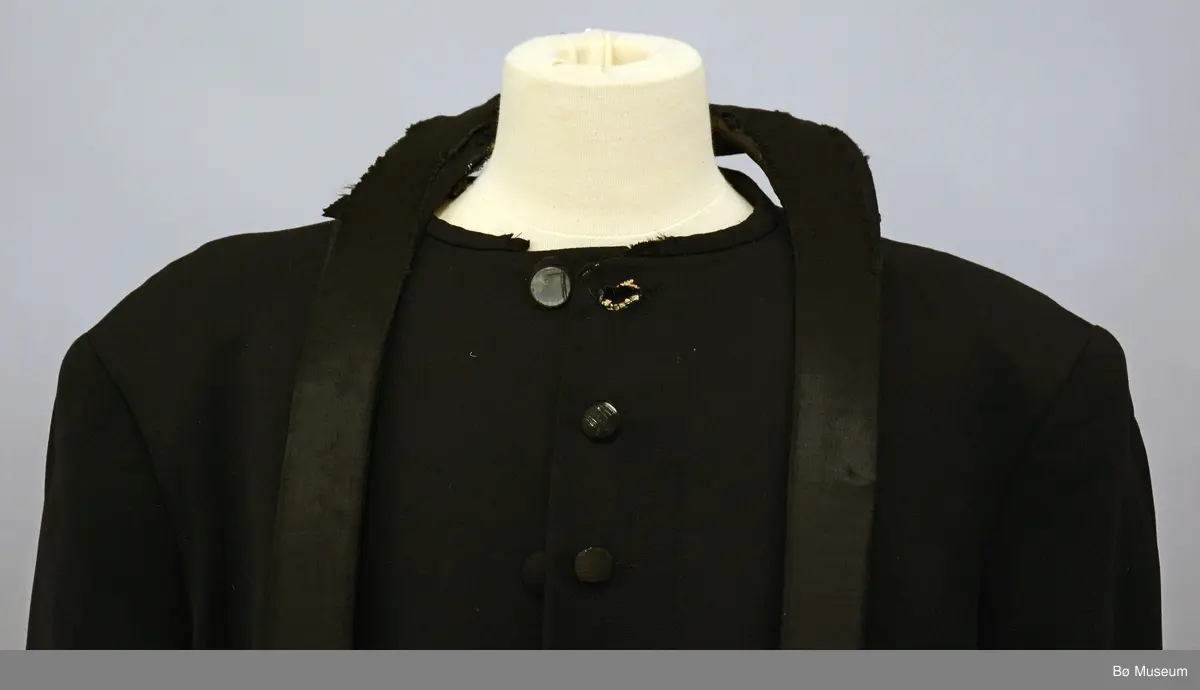 Svart fotsid samarie med tilhørende pipekrave (BØB.1995:0338.B). Drakten har langt bånd som er festet i kraven samt hvite mansjetter med hullsøm på ermene.

Fra registeringskort: Det lause bandet som går rundt nakken og ned på kvar side framme har stivt innlegg ned til liv et overtrekt med ullstoff. Frå livet og ned er fleire lag ull- stoff heile vegen overtrekt med svart sateng i ull og silke. Banda er feste med knappar til samarien, 5 på kvar side. Midt i nakken er det sydd fast til halslinninga. 21 knappar framme, trekte med silkesateng og markering av knapphol. Skjult knapping med 10 trekte knappar. Stor vidde i ryggen.