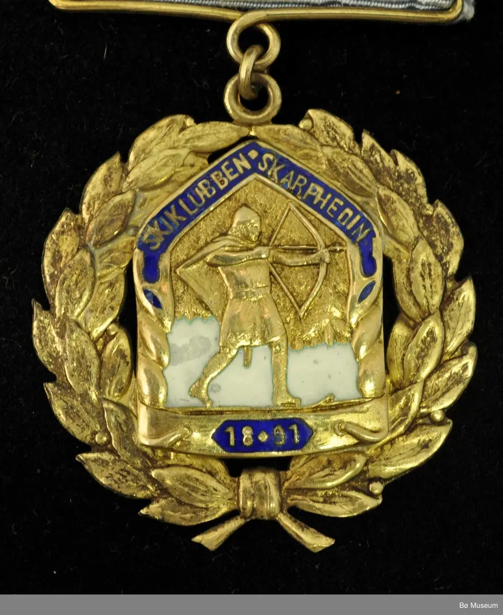Medalje med lyseblått bånd, navneplate og laurbærkrans rundt et merke med skigåer med pil og spent bue.