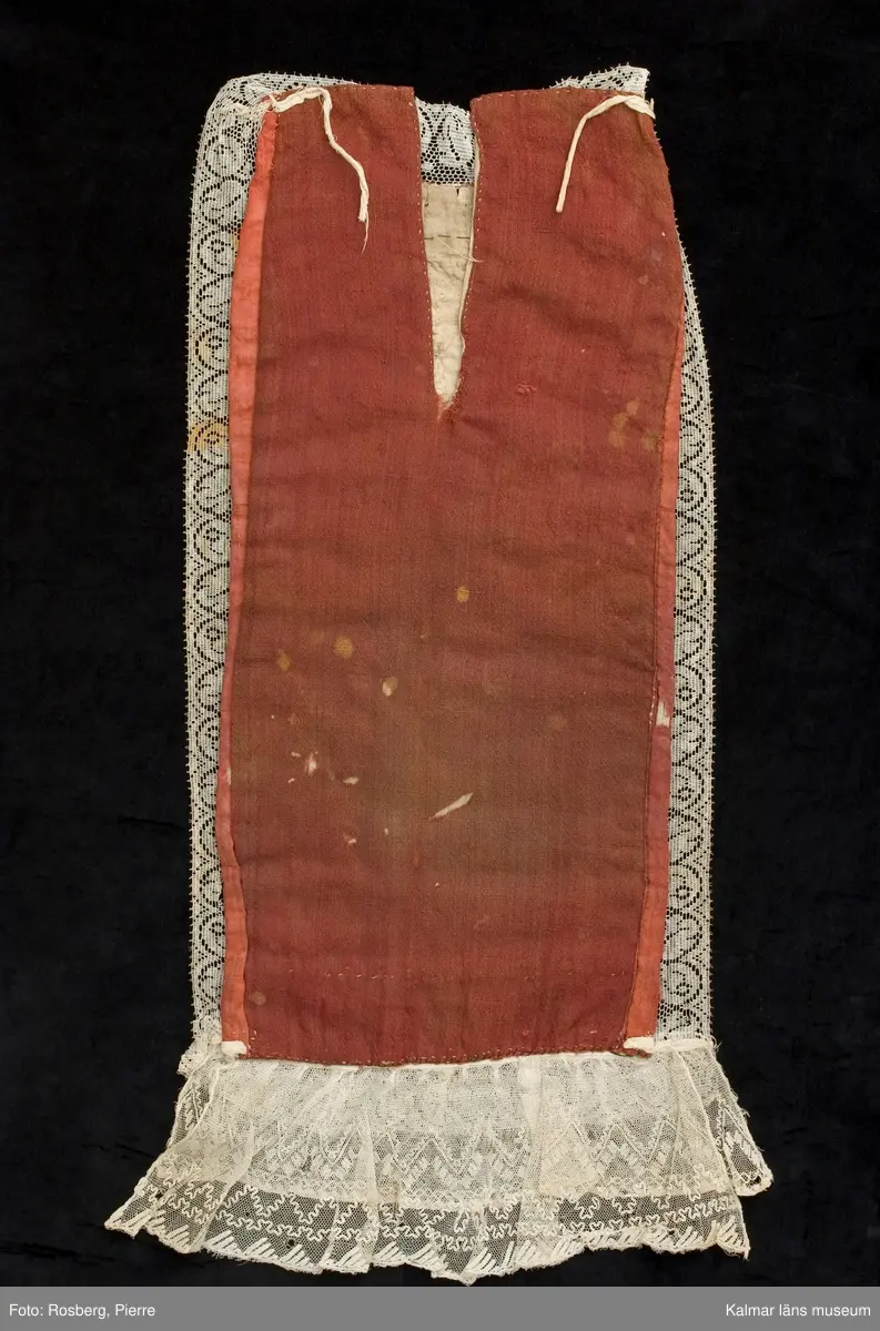 KLM 39363. Doppåse. 1800-tal. Sydd av rött, glättat ylletyg s.k. rask. Hela framsidan är broderad med små vita, svarta, gröna och ljus blå glaspärlor. Blommönster och girlanger. Ett smalt band av gulmetall är fastsytt ovanför pärlbroderierna, nära halsringningen. Halsringning, långsidor och nederkant är kantade med vit maskintillverkad spets. Vid nederkanten är två lager av vit broderad tyllspets fastsydd. Bakstycke av rödbrunt, tuskaftat ylletyg. Ett ca 19 cm långt sprund, från halshålet och ner, mitt bak. Knytband av linne fastsydda vid överkanten. Fodrad med vitt tuskaftat linne.