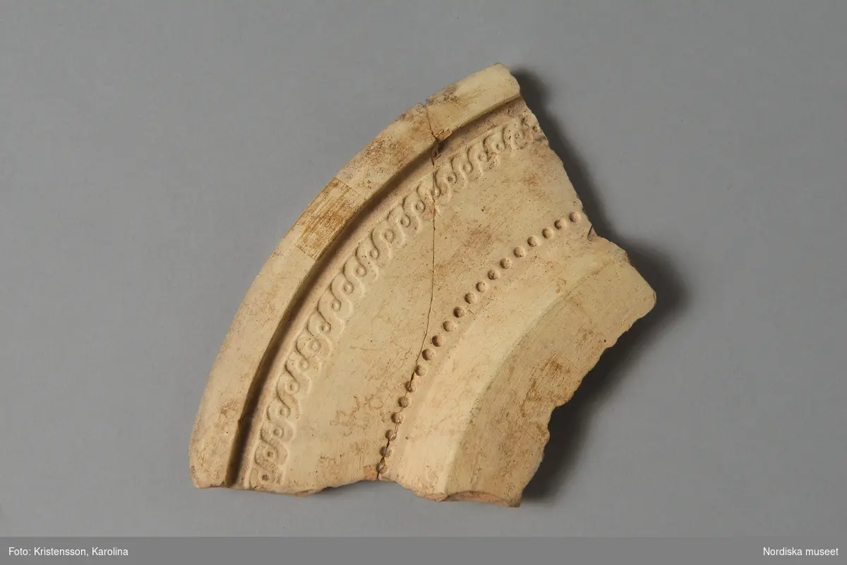 Huvudliggaren:
"Tallrik, fragment,  (lock?); flintgods, gjutet mönster  av s.k. bandflätning och punkteringar.  L ca 13 cm., br. ca 7,5 cm. Vänge-Gustafsberg". 

Del av tryckform till tallrik med mönster med bandfläta och punkteringar. Jordfynd från fabriken Vänge-Gustafsberg, Vänge sn, Uppland.
Sannolikt tillverkad 1785-1797 då Henrik Sten från Marieberg ledde tillverkningen.
/Tegnér 2008