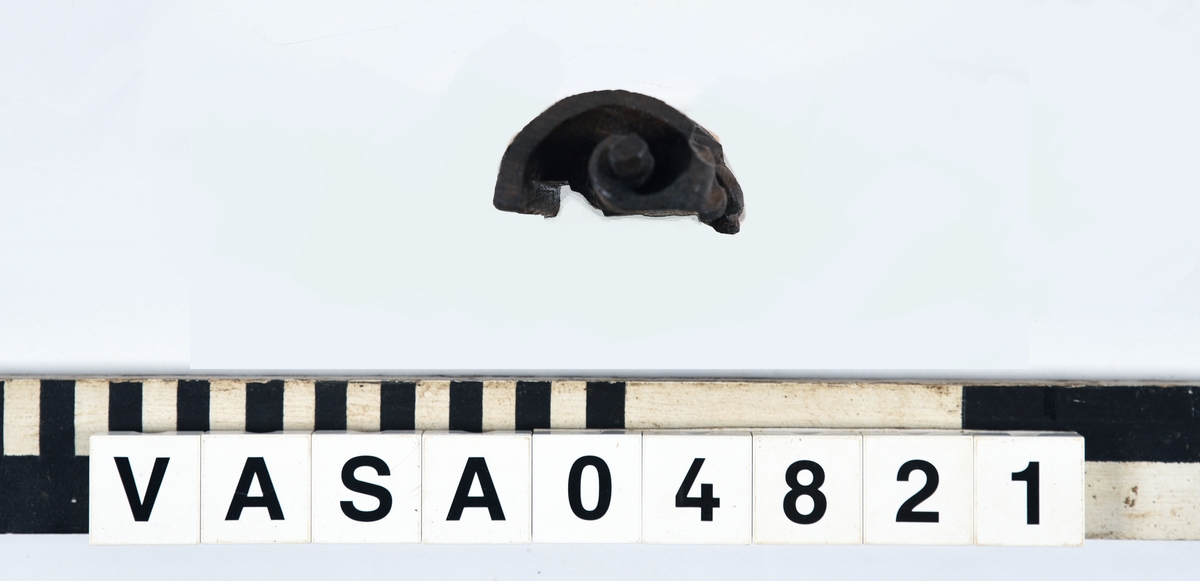 Rullverksornament.
Skulpterat litet, spänneliknande ornament, dekorerat i broskverksstil.
Det långsmala ornamentet är format som ett stycke kraftigt upprullad bit näver eller läder, med konkav bak- eller undersida. På baksidan är ornamentet profilerat för en list. Broskdekoren på ovansidan är formad som en maskaron.
Ornamentet är mycket välbevarat.

Text in English: A small, buckle-like ornament, decorated in a style similar to earshell work.
The long narrow ornament is formed as a piece of tightly rolled bark or leather, with a concave back or underside so as to match a batten or the like. The reverse side is profiled to fit a moulding. The earshell work on the upper side is shaped as a mask.
The ornament is well preserved.