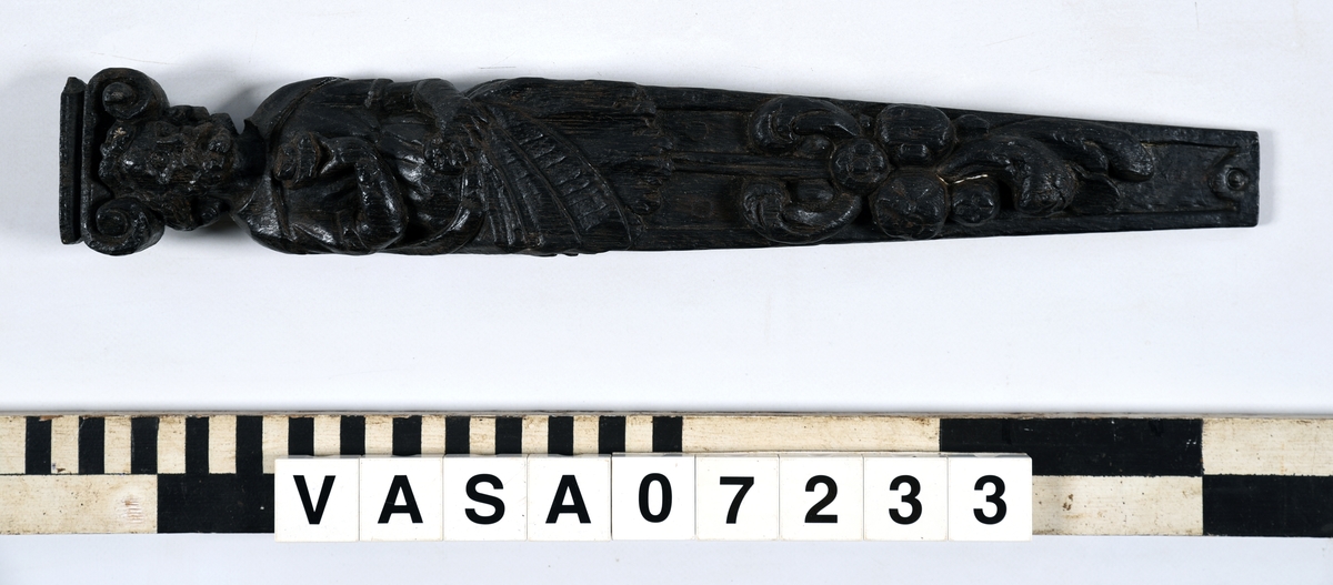Hermpilaster.
Liten skulpterad pilaster föreställande skäggig herm under joniskt kapitäl.
Huvudet är vridet mot vänster. Hermen är insvept i en mantel som hålls fast med vänster hand framför magen. I höger hand håller figuren ett okänt föremål (blomsterkälk?) framför bröstet. Pilasterskaftet är prytt med stora frukter och blad, fasthängande vid en stängel som delvis täcker det nedåt avsmalnande skaftets ramverk. Baksidan är slät.
Skulpturen är mycket välbevarad med sju spikhål.

Text in English: Small sculpted pilaster with a bearded Herm beneath an Ionic capital.
The head of the figure is turned to the left. The Herm is enveloped in a mantle, held fast with the left hand in front of its stomach. Its right hand holds an unknown object (a bunch of flowers?) in front of its breast. The shaft of the pilaster is decked with large fruit and leaves hanging from a stalk which partly cover the framing of the tapering shaft. Its back is flat.
The sculpture is very well preserved.
