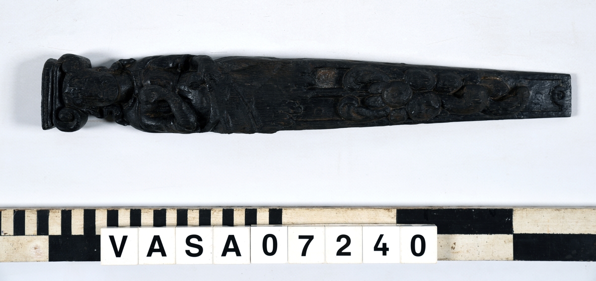 Hermpilaster.

Liten pilaster i form av en karyatid, med långt böljande hår under joniskt kapitäl.

Huvudet är vridet en aning åt höger. Med vänster hand hålls manteln samlad. I höger hand håller figuren en frukt eller dylikt framför bröstet. Pilasterskaftet är prytt med stora frukter och blad, fasthängande vid en stängel som delvis täcker det nedåt avsmalnande skaftets ramverk. Baksidan är slät.

Skulpturen är välbevarad, förutom ansiktet som är kraftigt slitet.

Fyra stycken spikhål, en övertäckt med en plugg.


Text in English: Sculpted small pilaster with a Caryatid with long wavy hair beneath an Ionic capital.

Its head is turned slightly to the right, its left hand holds its gathered mantle. Its right hand holds fruit or the like in front of its brests. The shaft of the pilaster is decorated with fruit and large leaves hanging from a stalk, which partly cover the framing of the tapering shaft. Its back is flat.

The sculpture is somewhat worn, its facial expression has been obliterated.