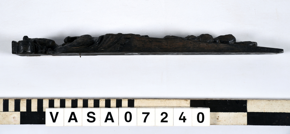 Hermpilaster.

Liten pilaster i form av en karyatid, med långt böljande hår under joniskt kapitäl.

Huvudet är vridet en aning åt höger. Med vänster hand hålls manteln samlad. I höger hand håller figuren en frukt eller dylikt framför bröstet. Pilasterskaftet är prytt med stora frukter och blad, fasthängande vid en stängel som delvis täcker det nedåt avsmalnande skaftets ramverk. Baksidan är slät.

Skulpturen är välbevarad, förutom ansiktet som är kraftigt slitet.

Fyra stycken spikhål, en övertäckt med en plugg.


Text in English: Sculpted small pilaster with a Caryatid with long wavy hair beneath an Ionic capital.

Its head is turned slightly to the right, its left hand holds its gathered mantle. Its right hand holds fruit or the like in front of its brests. The shaft of the pilaster is decorated with fruit and large leaves hanging from a stalk, which partly cover the framing of the tapering shaft. Its back is flat.

The sculpture is somewhat worn, its facial expression has been obliterated.