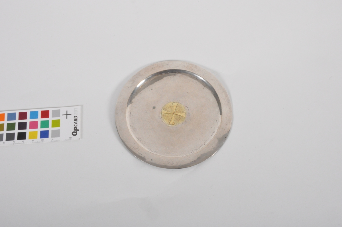 Oblatdisk i sølv. I diskens senter er et gullforgylt pattéekors i en sirkel risset inn. På diskens bakside er et våpenskjold risset inn. Trolig generalmajor Hausmanns. Se HM.00544.