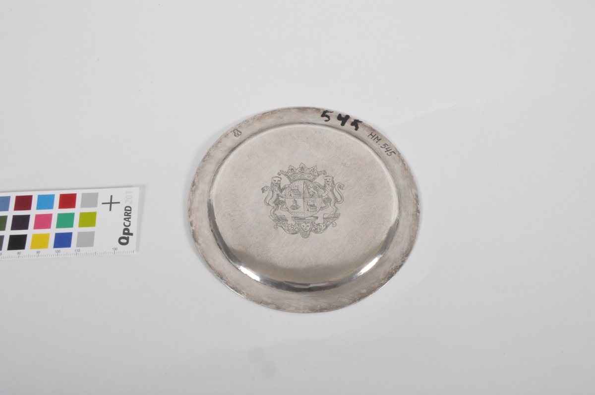 Oblatdisk i sølv. I diskens senter er et gullforgylt pattéekors i en sirkel risset inn. På diskens bakside er et våpenskjold risset inn. Trolig generalmajor Hausmanns. Se HM.00544.