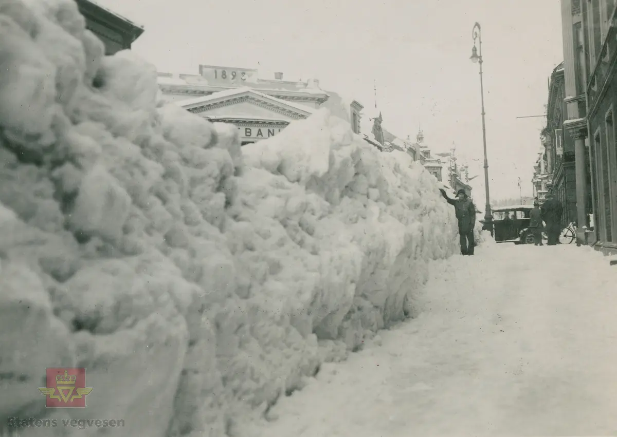 Fra hovedveiene rundt i Vest-Agder vinteren 1937 i følge merking i album, og med store snømengder. I  bakgrunnen på dette bildet vises Norges Bank 1893. Ref. til "Meddelelser fra Veidirektøren", Nr. 3-1937. "De vanskelige sneforhold på Sørlandet i år". Av avdelingsingeniør T. Backer. Se vedlegg i Nedlastinger.