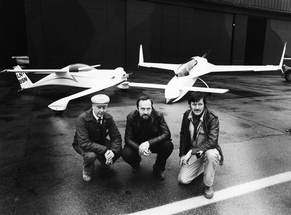 Lufthavn/Flyplass. To hjemmebyggede fly, det ene LN-BSN, parkert ved en hangar. Tre personer, eiere av flyene, sitter foran.