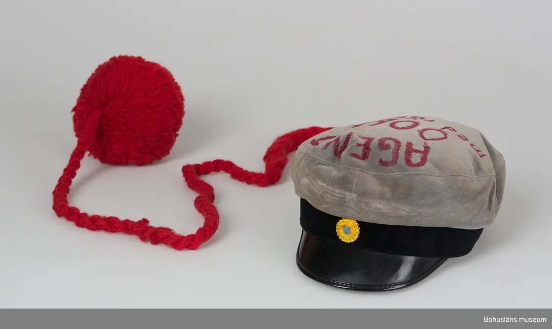 Grå realskolemössa med kulle av sammet och foder av gult och blått siden. Märkt med etikett "Gergils".
Nedhängande snodd av tvinnat tvåslaget med tofs i änden och påskrift i rött på kullen med texten:
 "AGENT 
0066
med rätt att
ta
STUDENT
EN