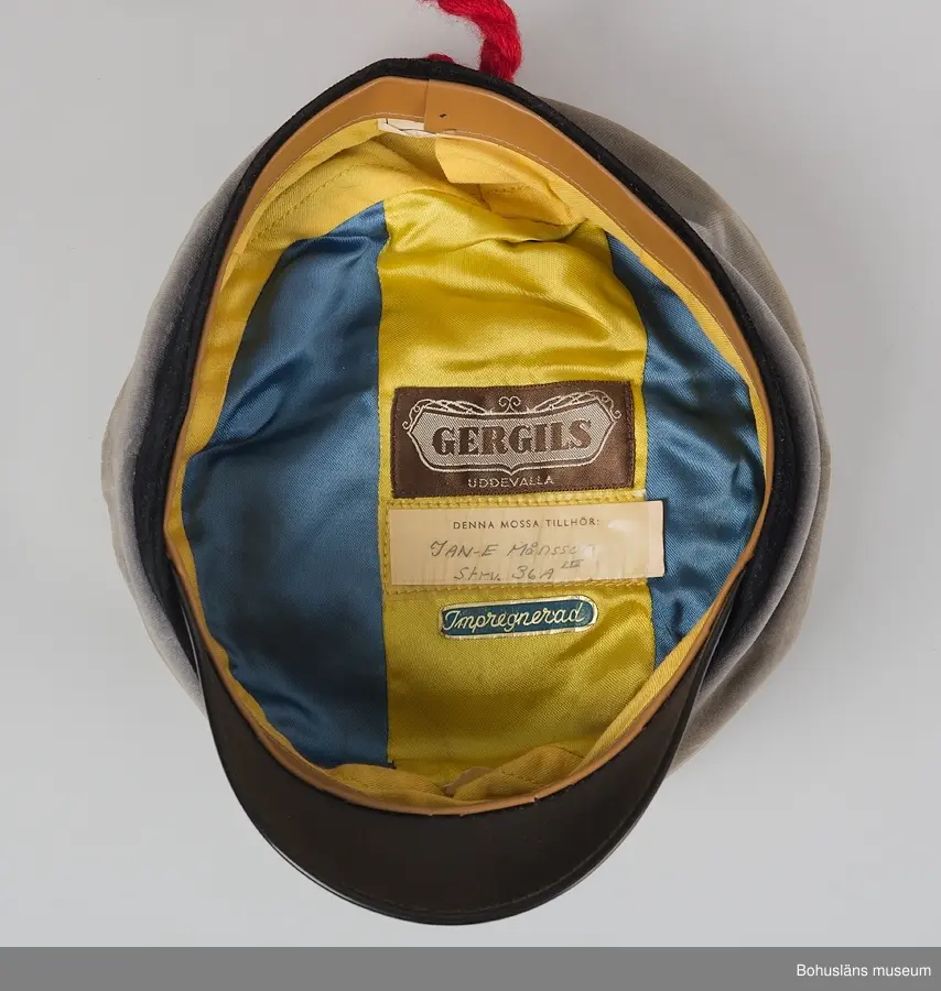 Grå realskolemössa med kulle av sammet och foder av gult och blått siden. Märkt med etikett "Gergils".
Nedhängande snodd av tvinnat tvåslaget med tofs i änden och påskrift i rött på kullen med texten:
 "AGENT 
0066
med rätt att
ta
STUDENT
EN