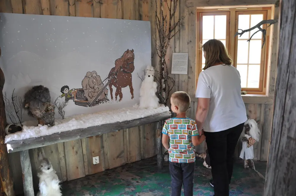 Inne i trollhuset står ett barn og en voksen og ser på et maleri. Maleriet er av en unge som står bak en slede med to voksne som blir dratt av en hest.