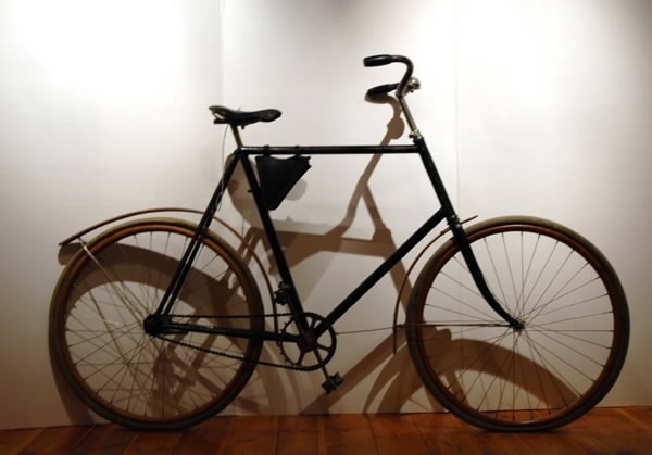 Cykel av stålrör. Diamond-ram. Lädersadel på metallbotten och stavfjädrar. Vertygsväska av läder i remmar under övre ramröret. Styre med handtag av konstmassa. Ringklocka. Hjul av träfälgar på suspensionsekrar. Stänkskärmar av trä. Kedja med länkar av 1,9 cm längd omväxlande med länkar av 2,4 cm längd och förenade medelst rullar. Svart med förkromat styre, brungula fälgar och stänkskärmar.