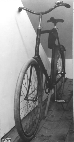 Cykel av stålrör. Diamond-ram. Lädersadel på metallbotten och stavfjädrar. Vertygsväska av läder i remmar under övre ramröret. Styre med handtag av konstmassa. Ringklocka. Hjul av träfälgar på suspensionsekrar. Stänkskärmar av trä. Kedja med länkar av 1,9 cm längd omväxlande med länkar av 2,4 cm längd och förenade medelst rullar. Svart med förkromat styre, brungula fälgar och stänkskärmar.