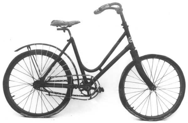 Cykel, flickstorlek av stålrör. Bakskärm och fälgar av trä. Stoppad lädersadel. Styre med korkhandtag och ringklocka. På bakskärmen hål för hjulskydd. Kedjeskydd av trä med hål för nät eller dylikt. Kedja av 2,4 cm långa länksidor, förenade genom massiva mellanstycken. Pedaler av gummi. Luftfyllda gummidäck. Ekrarna sammanlödda två och två vid navet. På styrrörets framsida firmamärke, se bild. Cykeln saknar broms och frihjul.