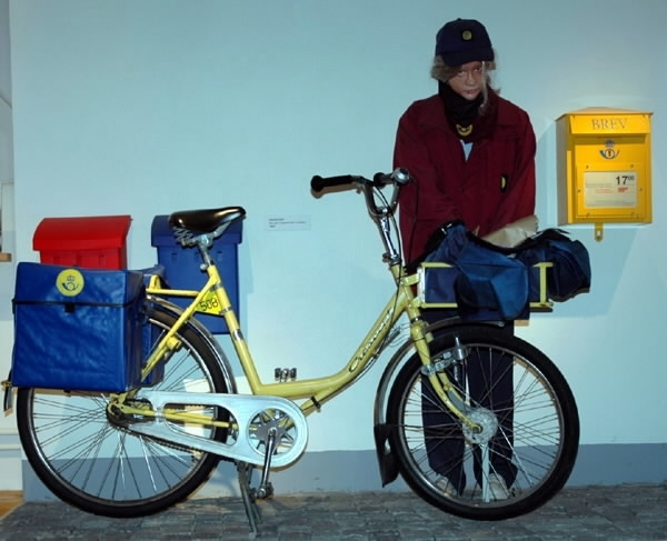 Gul postcykel (a) med svarta handtag och svart sadel. En mörkt grön-blå väska (b) av textil fram med postens logotype. 
Mått b: längd 50 cm, bredd 68 cm, höjd 28 cm. Samt två blå plastväskor bak (c-d). Mått c-d: längd 33 cm, bredd 24 cm, höjd 35 cm.