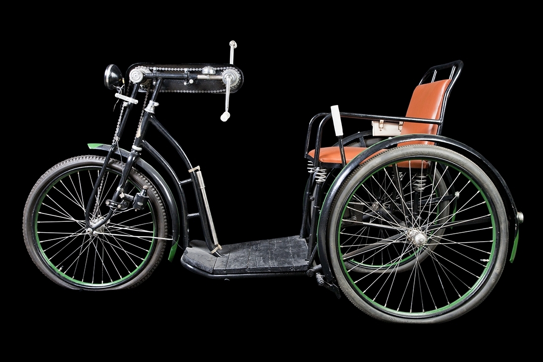 Trehjulscykel, rullstolscykel, svart med röd/brun sits i skinnimitation. Tre hjul som drivs med handvev genom en kedja till framhjulet. Handbroms.  Reglerbart ryggstöd. Fotplatta av trä, svart. Vit börs eller väska på sidan av sitsen.